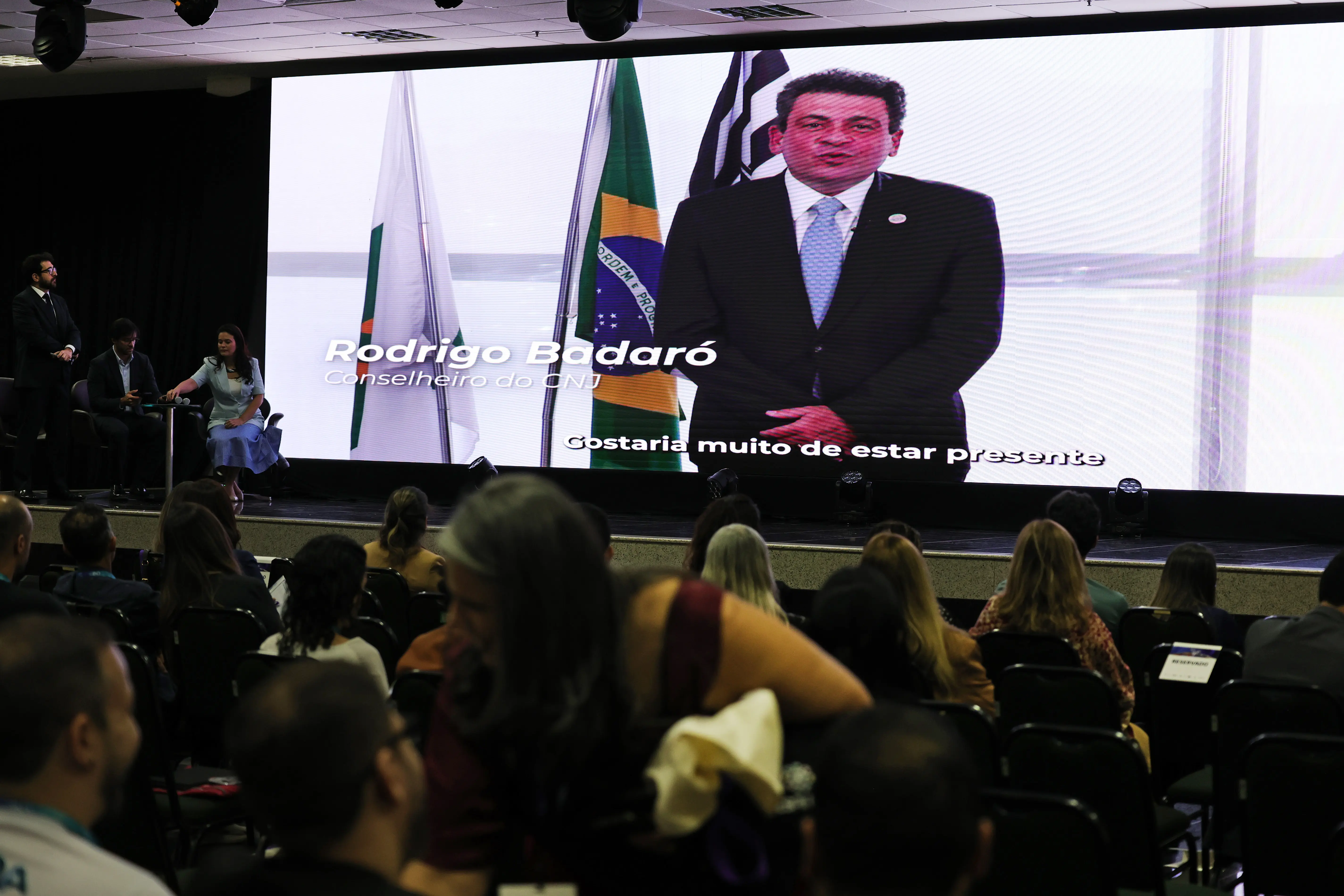 Telão exibe participação virtual do conselheiro do CNJ Rodrigo Badaró, que discursa ao lado das bandeiras nacional e estadual; público acompanha sentado no auditório.