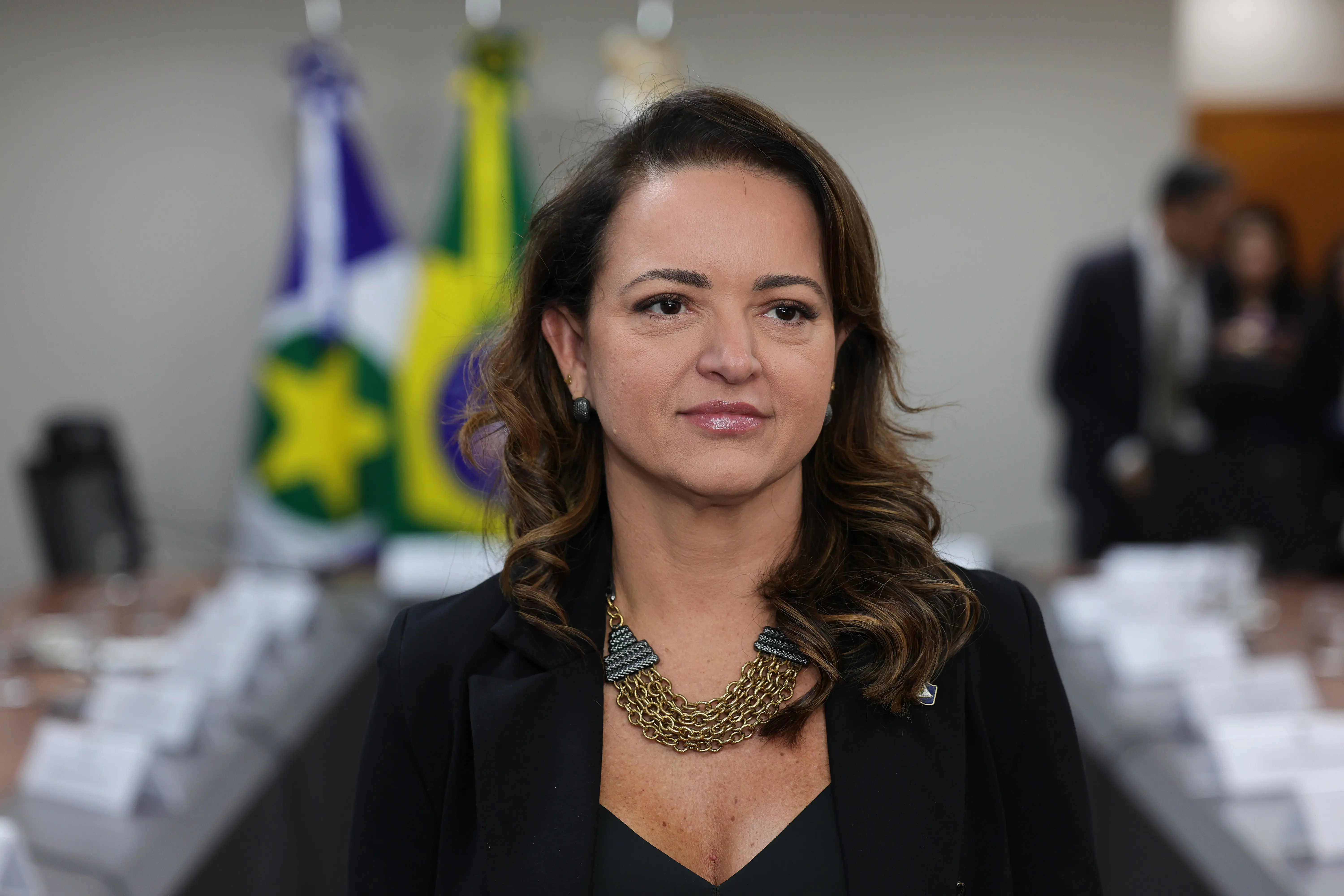 Primeiro plano de uma mulher de cabelos castanhos ondulados, usando blazer preto e colar dourado de correntes. Ao fundo desfocado, aparecem a mesa de reuniões, bandeiras e outras pessoas no ambiente.