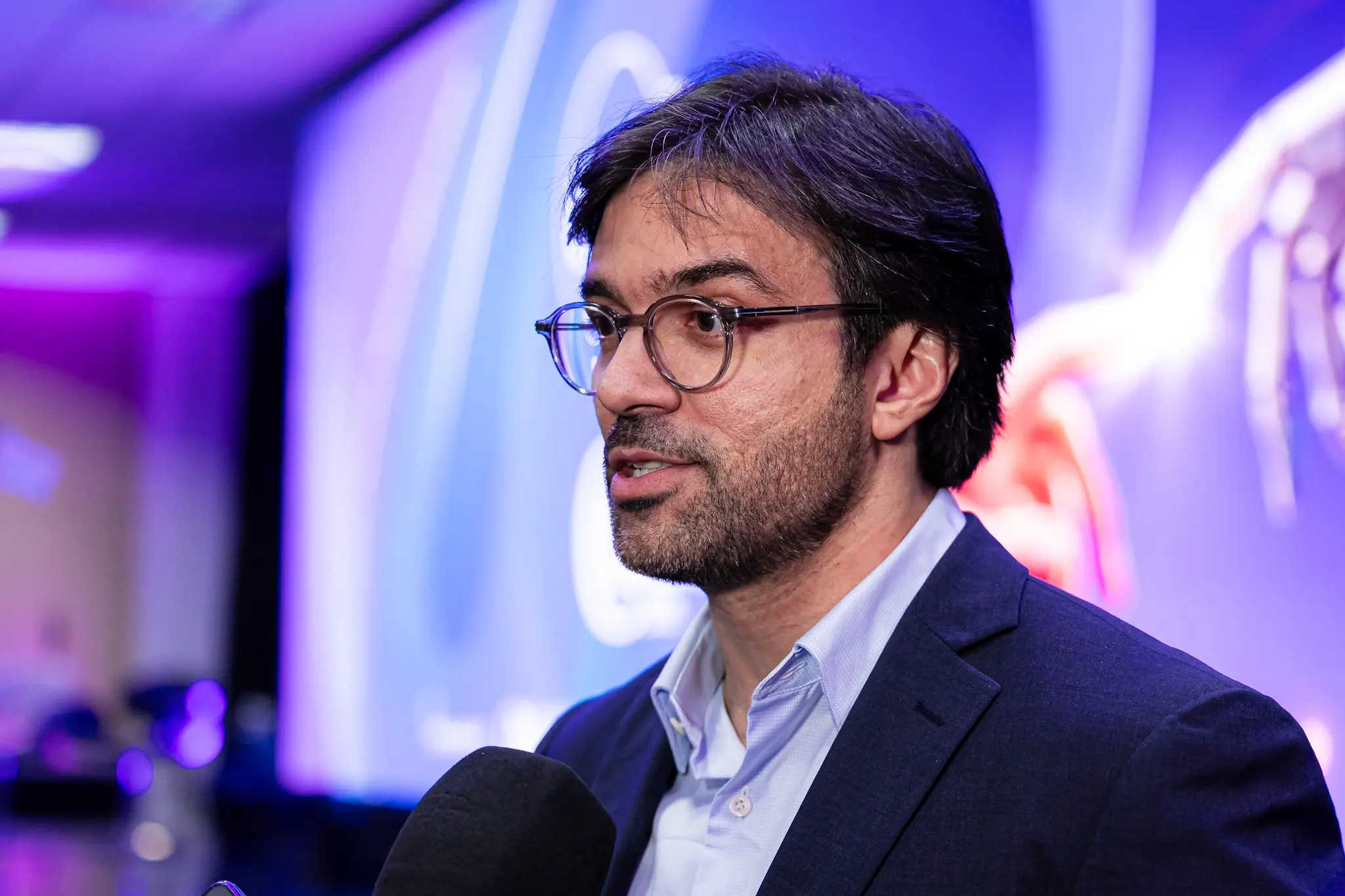 Juiz Jeremias fala próximo ao microfone, de óculos e blazer azul-marinho, com fundo em tons de roxo e azul iluminado.