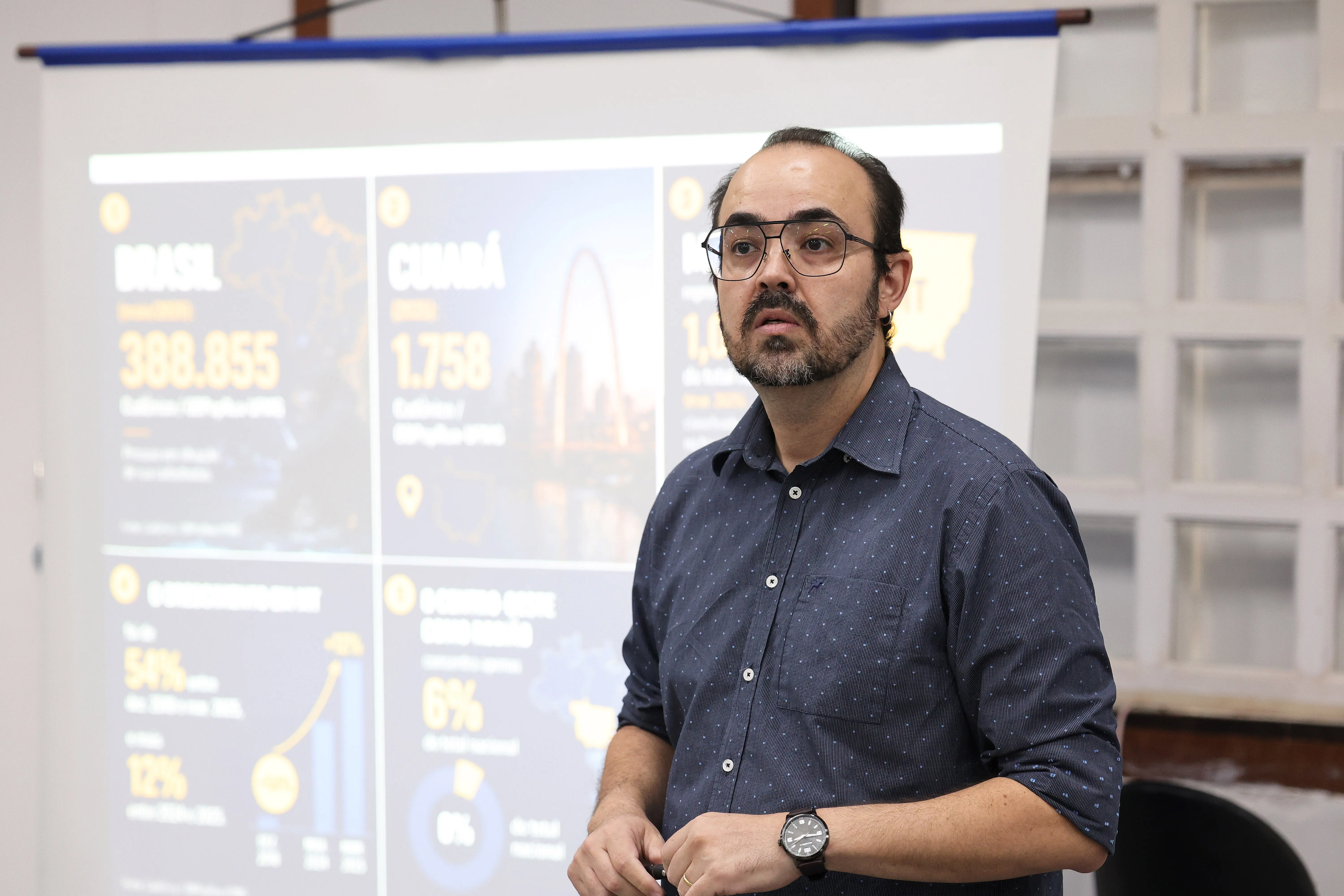 Homem de óculos, barba grisalha e camisa azul-marinho discursa em pé diante de telão com dados estatísticos sobre população em situação de rua no Brasil e em Cuiabá.