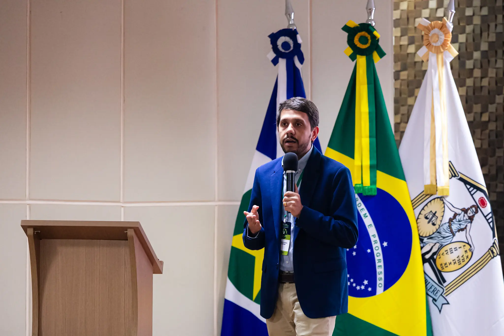 Homem de cabelos escuros e barba, blazer azul e calça bege, discursa ao microfone com crachá, bandeiras de MT, Brasil e do TJMT ao fundo.