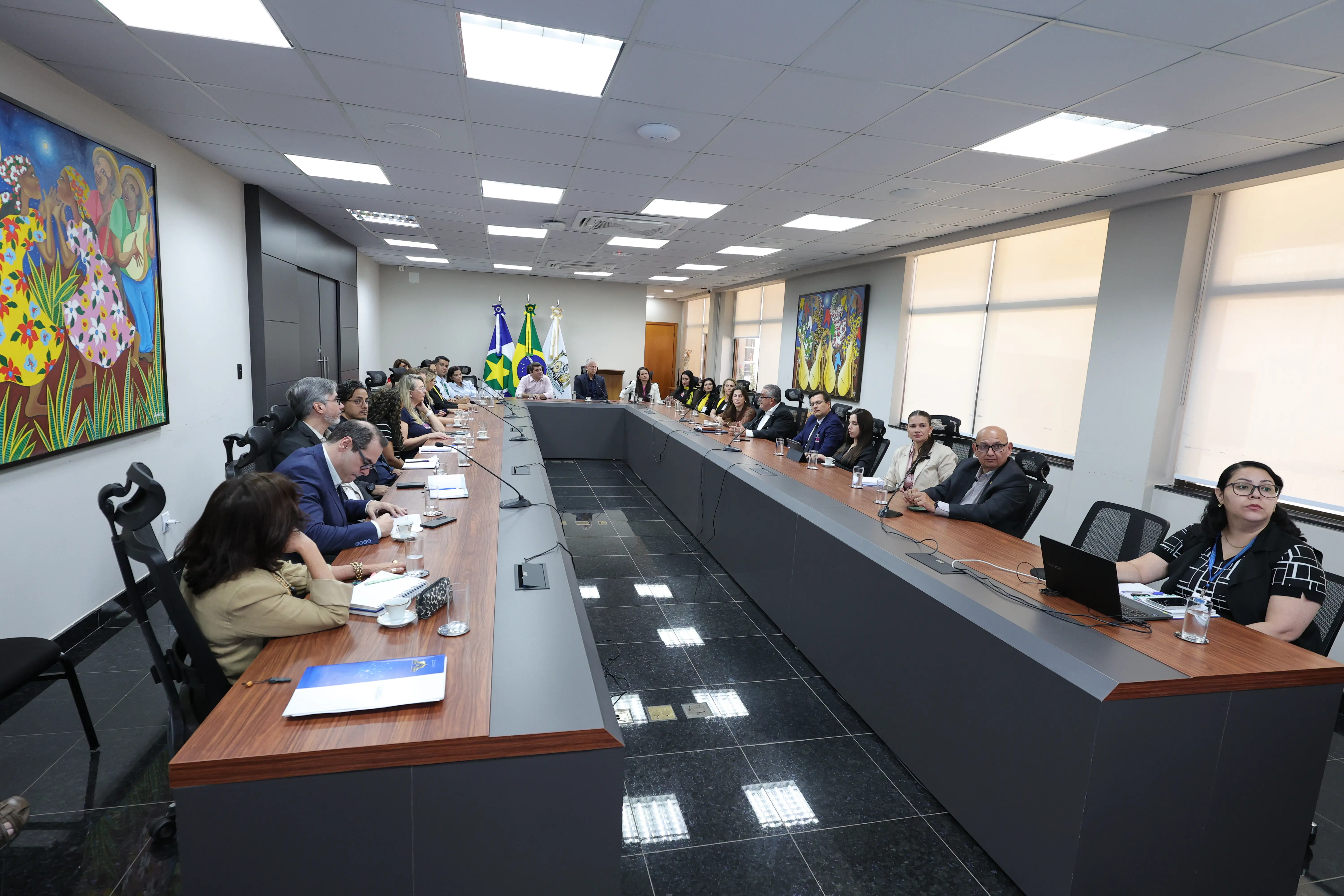 Autoridades e servidores em reunião de mesa em U no TJMT, com bandeiras do Brasil, Mato Grosso e do Tribunal ao fundo, entre painéis artísticos coloridos na parede.