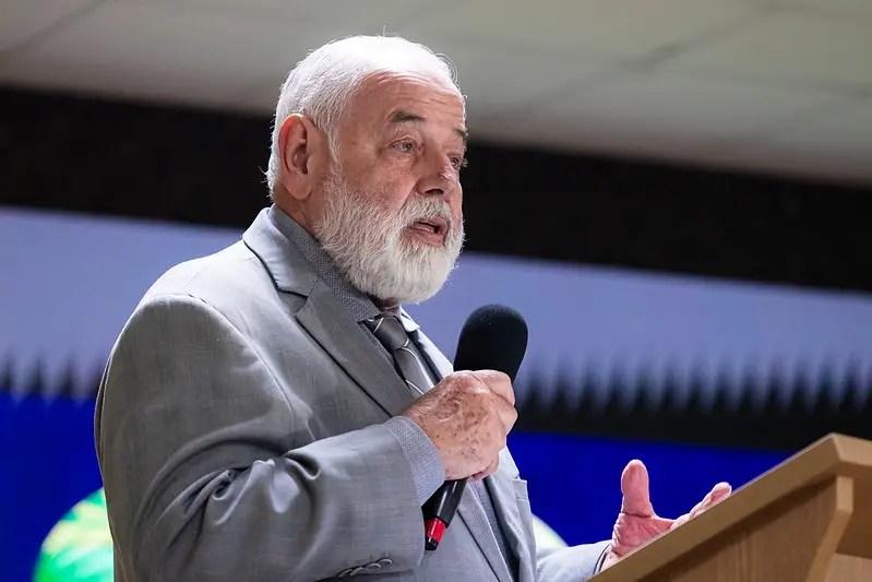 Foto horizontal que mostra o desembargador José Zuquim Nogueira do busto pra cima, falando ao microfone em um púlpito de madeira. ele é um senhor branco, de cabelo e barba brancos, olhos claros, usando terno e gravata cinza claro.
