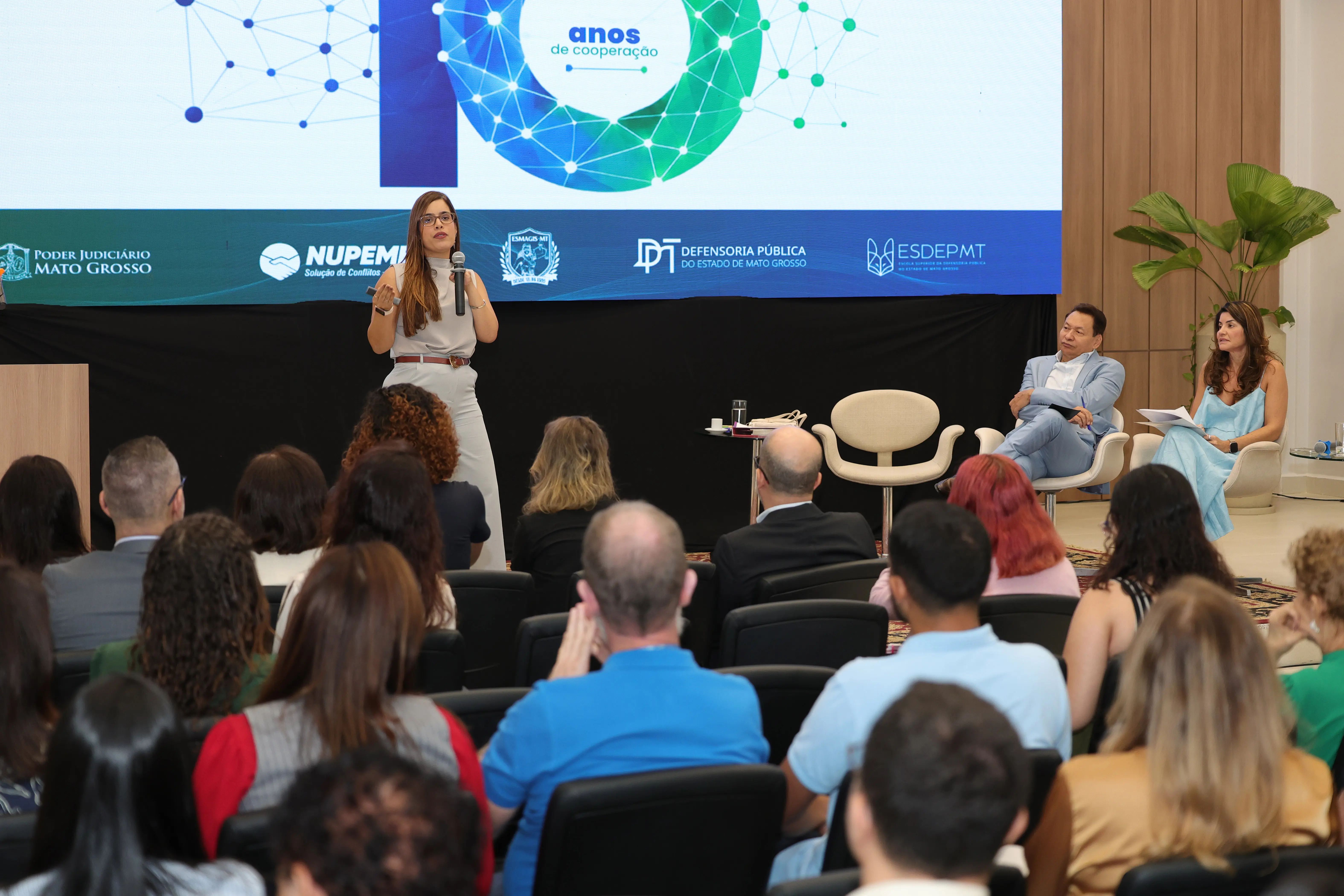 Visão geral de um evento em um auditório. Uma mulher fala ao microfone em pé no palco, que tem uma grande tela ao fundo com vários logotipos e a frase "anos de cooperação". Um homem de terno azul e uma mulher de vestido azul claro estão sentados à direita do palco. Várias pessoas estão sentadas na p