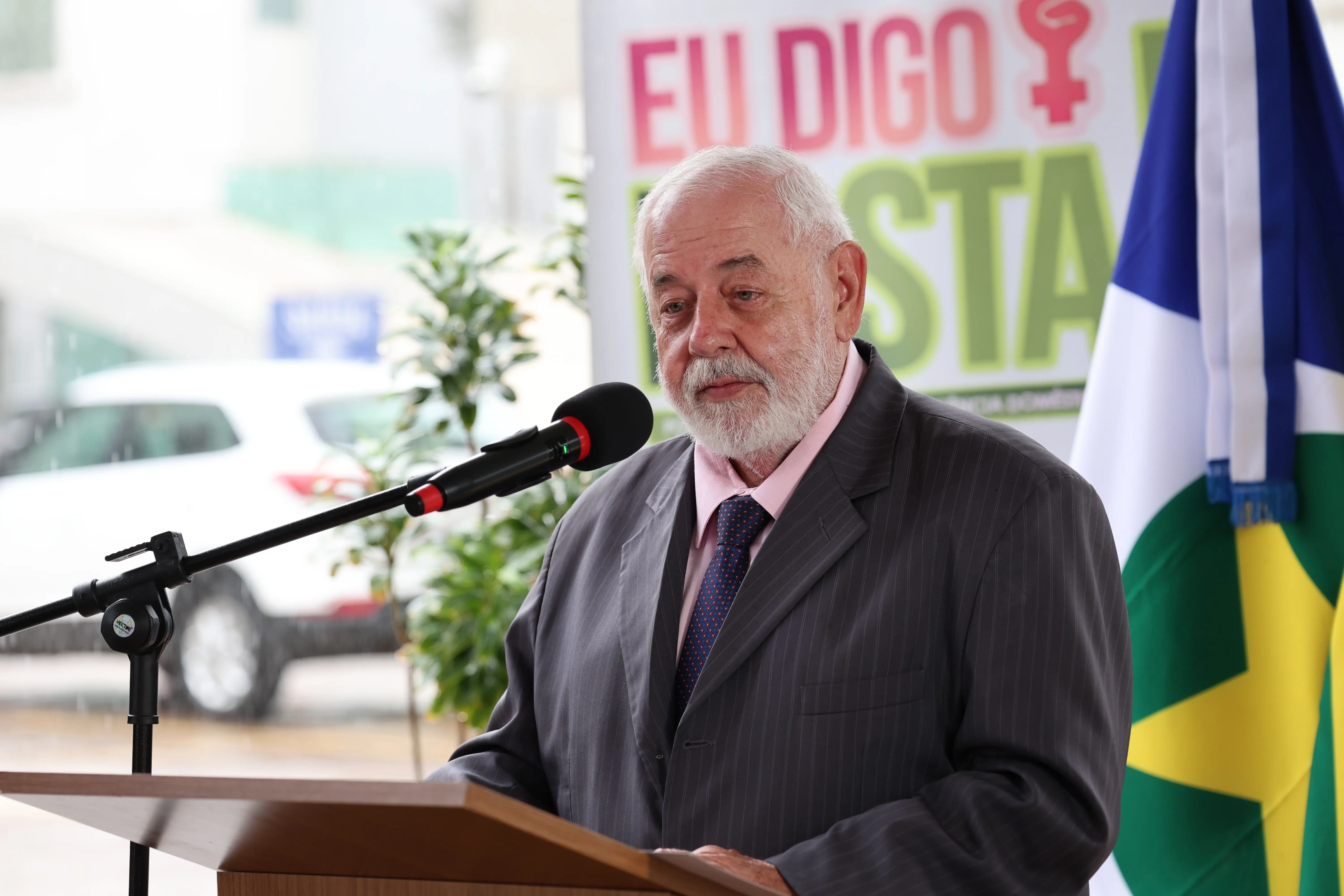 José Zuquim Nogueira, homem de pele clara, barba e cabelos brancos. Veste terno cinza e gravata azul. Está sério, atrás de um púlpito com microfone. Ao fundo, banner com a frase 