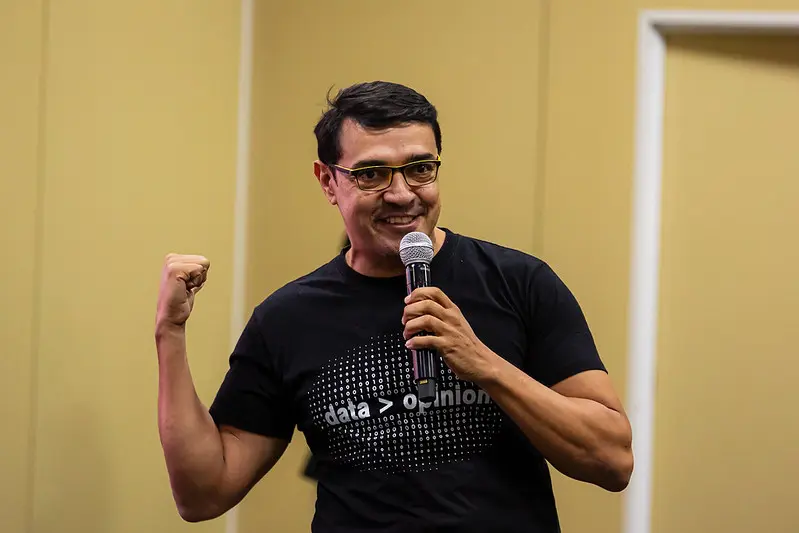 Foto horizontal que mostra Welkey Costa, coordenador do laboratório de inovação do Tribunal de Justiça do Ceará, da cintura pra cima, falando ao microfone e gesticulando. ele é um homem alto, pardo, de cabelo liso e preto, usando camiseta preta estampada e óculos de grau.