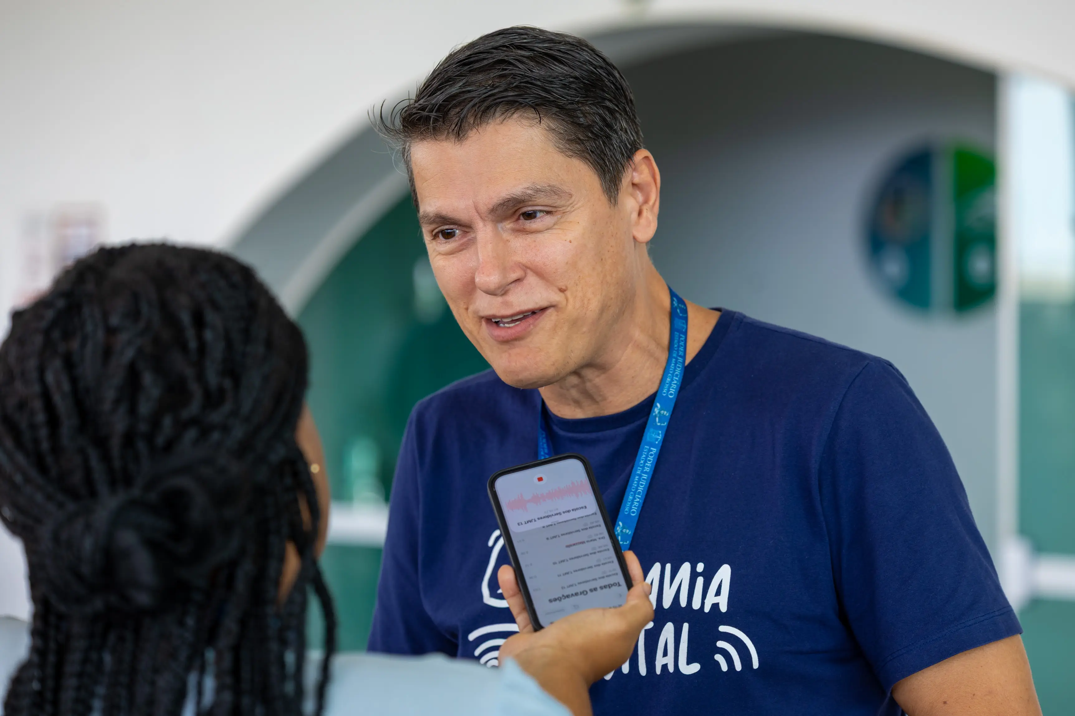 Homem branco, de camiseta azul com crachá falando ao microfone de um celular durante entrevista.
