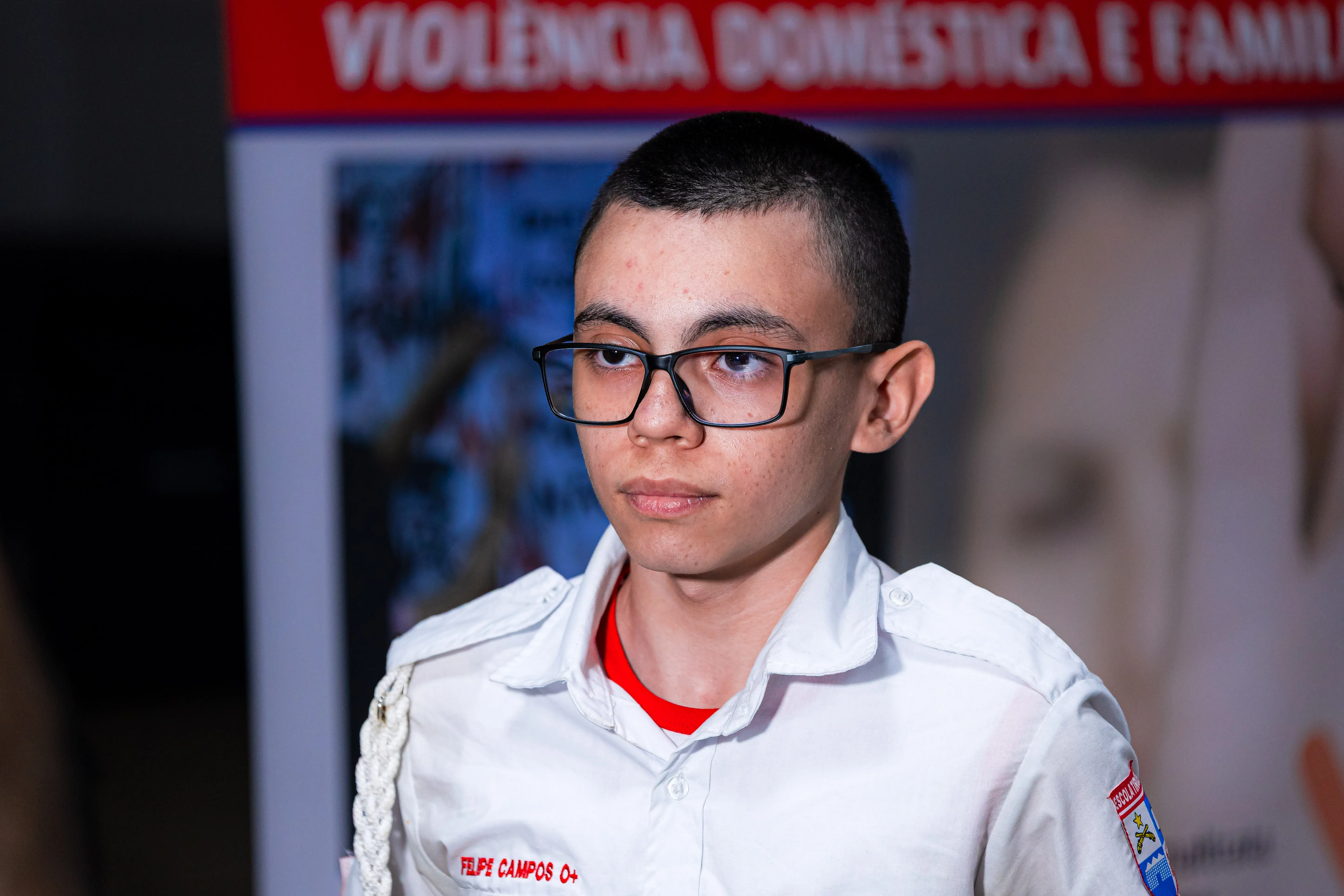 Jovem de óculos e uniforme escolar branco, com identificação no bolso, observa em frente a banner vermelho sobre violência doméstica e familiar.