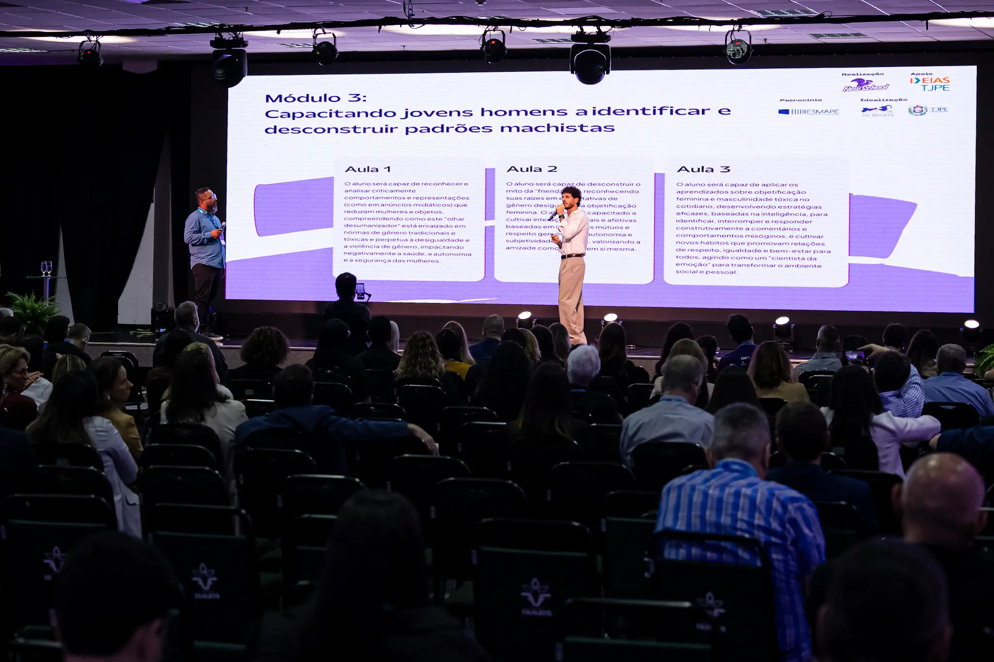 Palco de auditório visto de trás da plateia. No centro, um homem com camisa social branca e calça clara fala ao microfone. No telão, lê-se "Módulo 3: Capacitando jovens homens a identificar e desconstruir padrões machistas" com três caixas de texto abaixo.