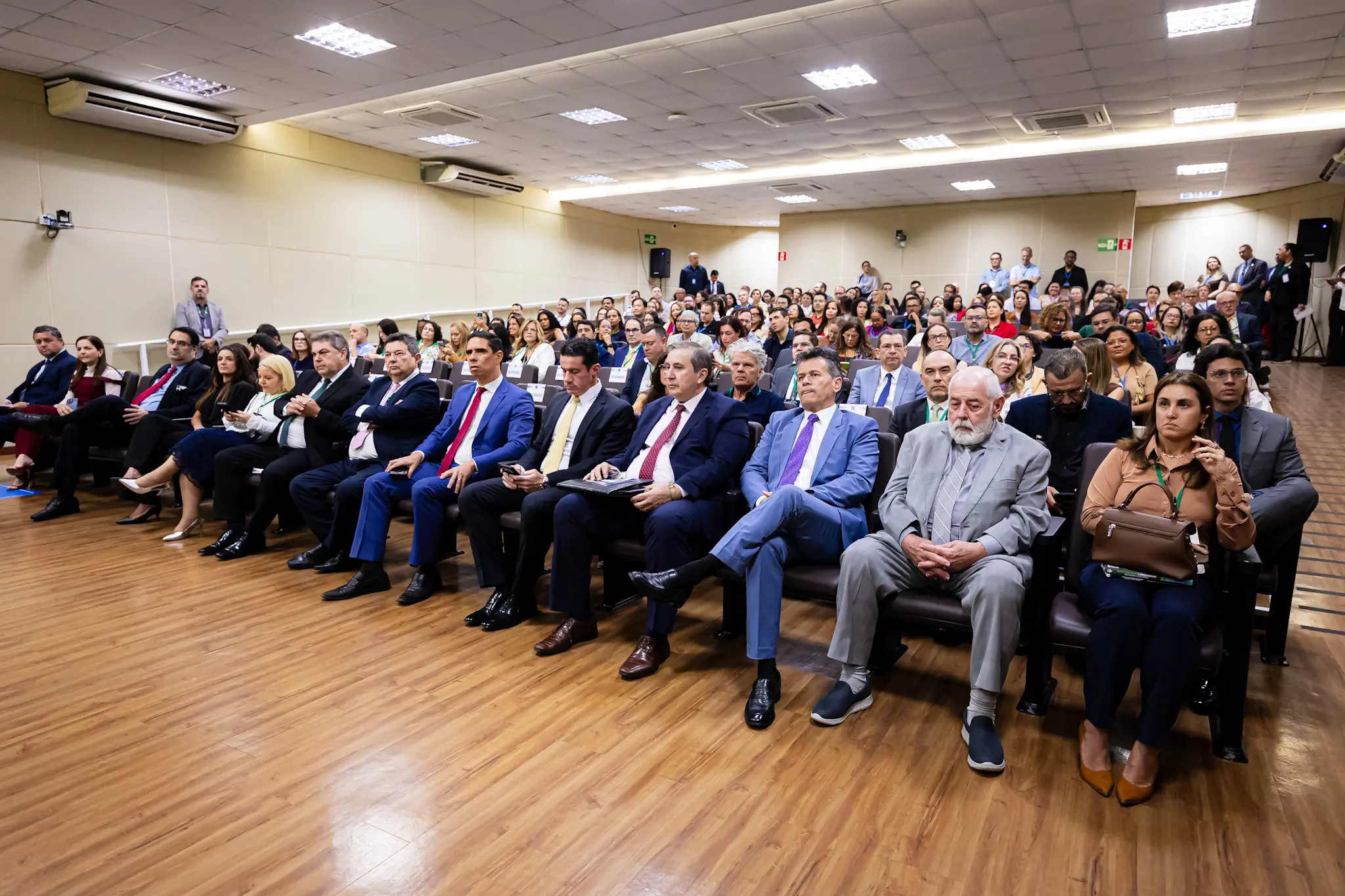 Visão geral do público sentado em poltronas de um auditório com piso de madeira. A maioria veste trajes formais, como ternos e vestidos. Na primeira fileira, destacam-se homens de terno e gravata e uma mulher com bolsa marrom.