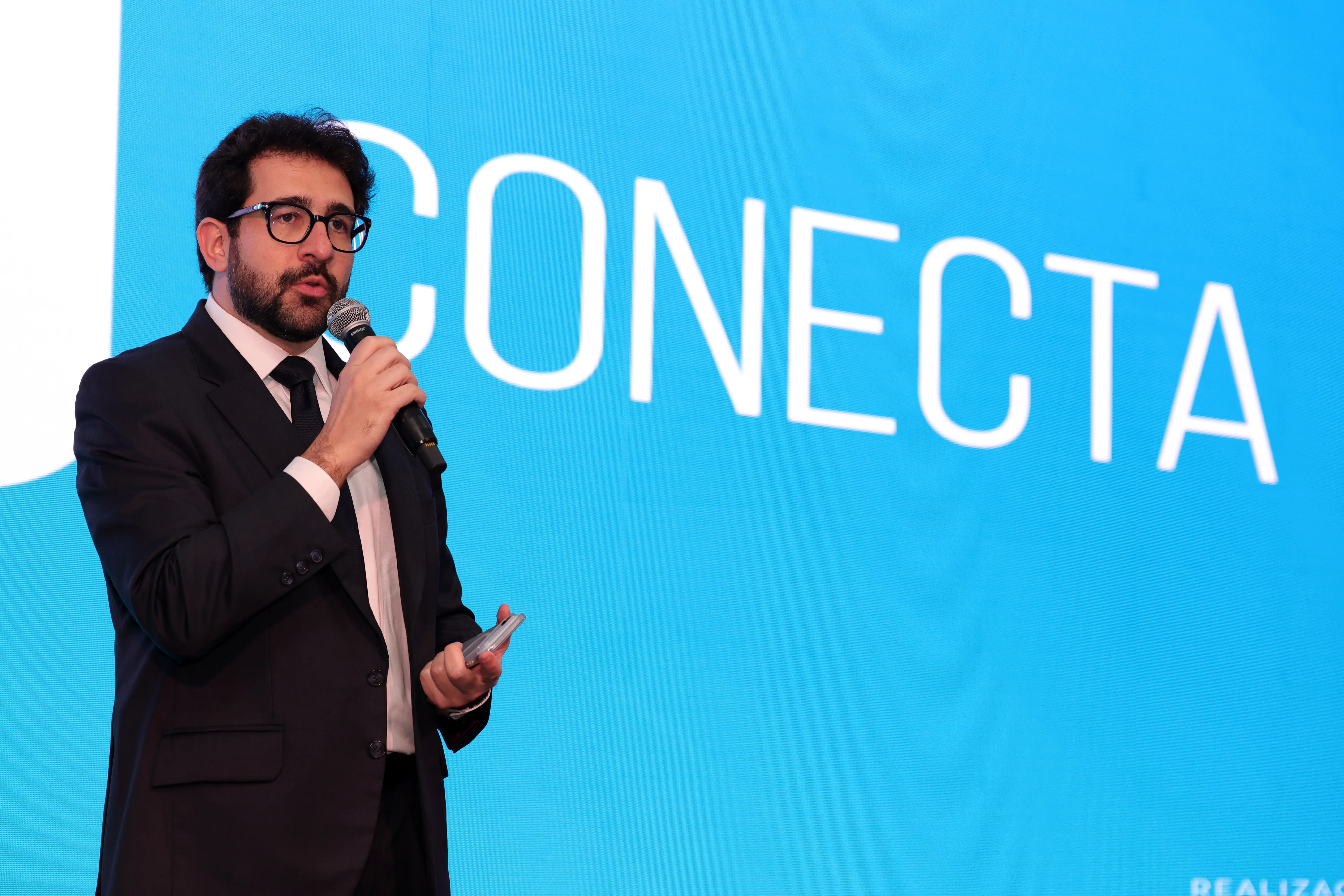 Juiz Henrique Paiva discursa ao microfone, de óculos, terno e gravata pretos, diante de telão azul com a palavra "Conecta" ao fundo.