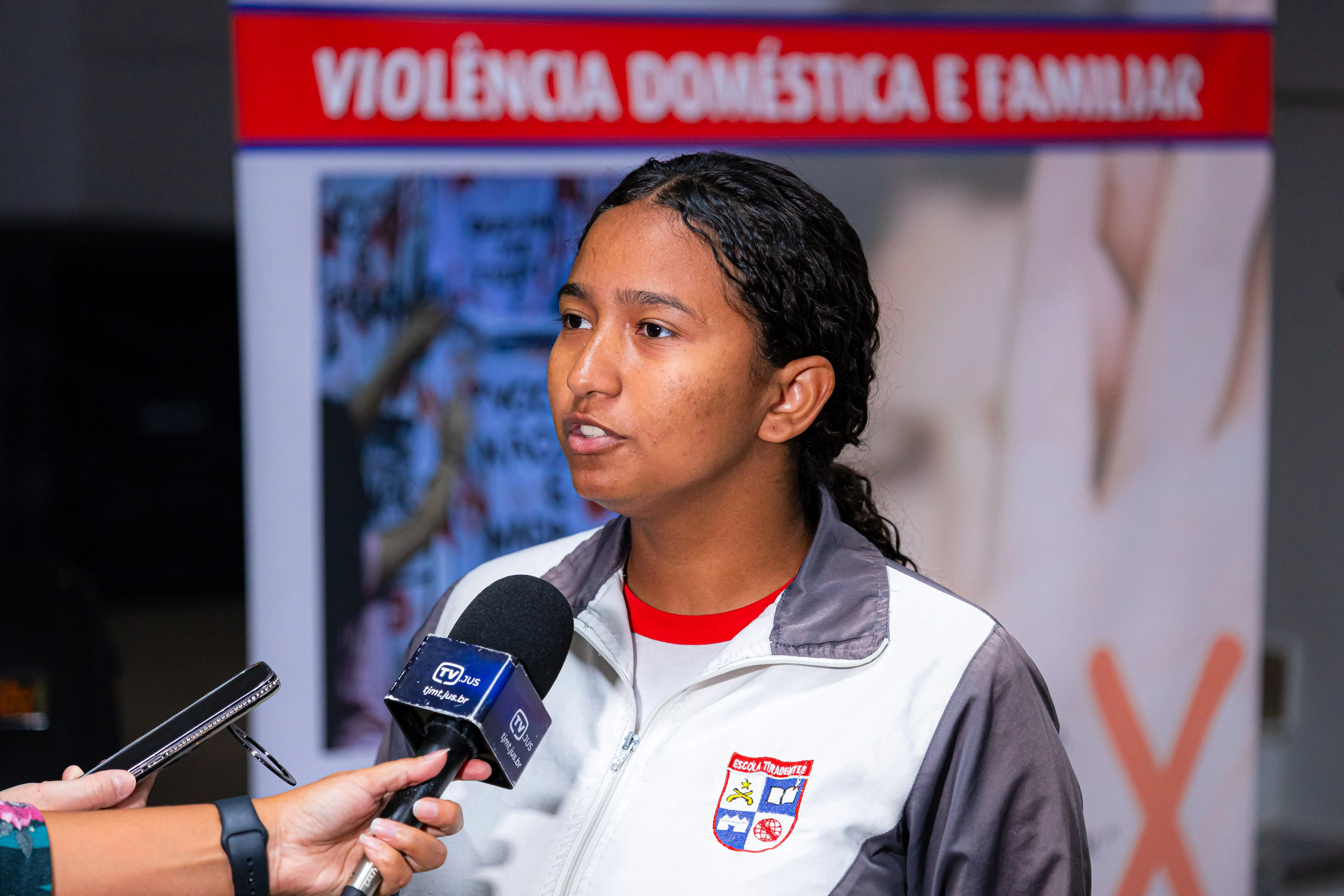 Jovem de cabelos cacheados e jaqueta da Escola Tiradentes é entrevistada por microfone da TV Jus, diante de banner sobre violência doméstica e familiar.