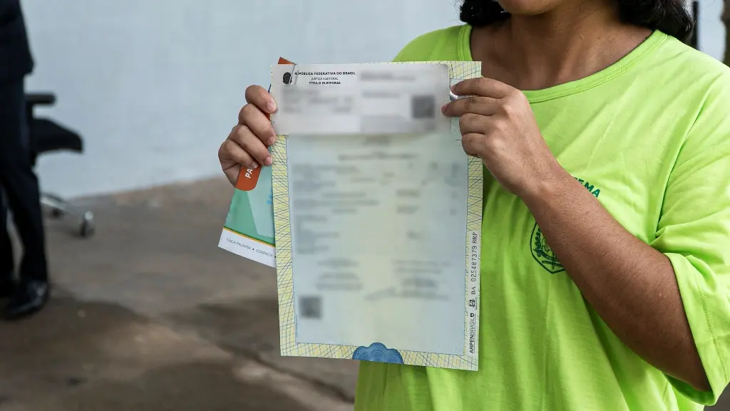Foto horizontal que mostra uma adolescente em medida socioeducativa segurando seu documento, que está com as informações borradas. Ela tem pele negra e usa camiseta verde neon do centro socioeducativo.