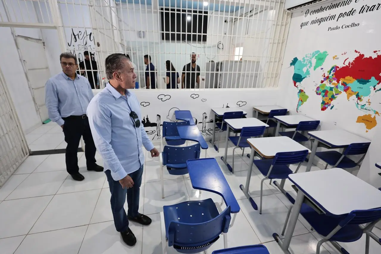 Dois homens em uma sala de aula com paredes brancas. À direita, um mapa-múndi colorido e carteiras escolares azuis. Ao fundo, uma grade branca separa o ambiente de um corredor onde há outras pessoas.