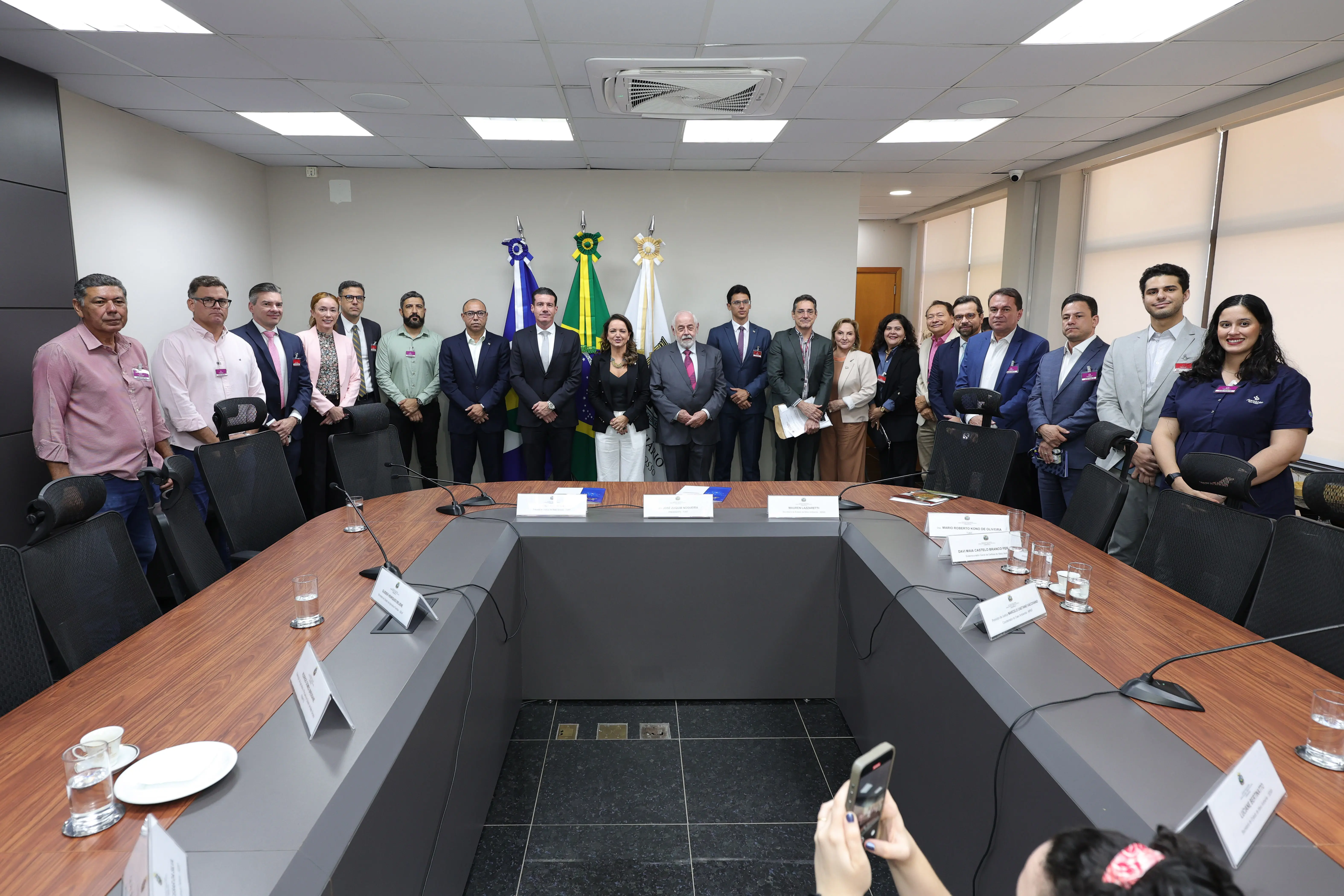 Plano geral exibindo um grupo de homens e mulheres alinhados em pé ao fundo de uma sala de reuniões, atrás da mesa retangular. Ao centro do grupo, estão dispostas as bandeiras do Brasil e de Mato Grosso.