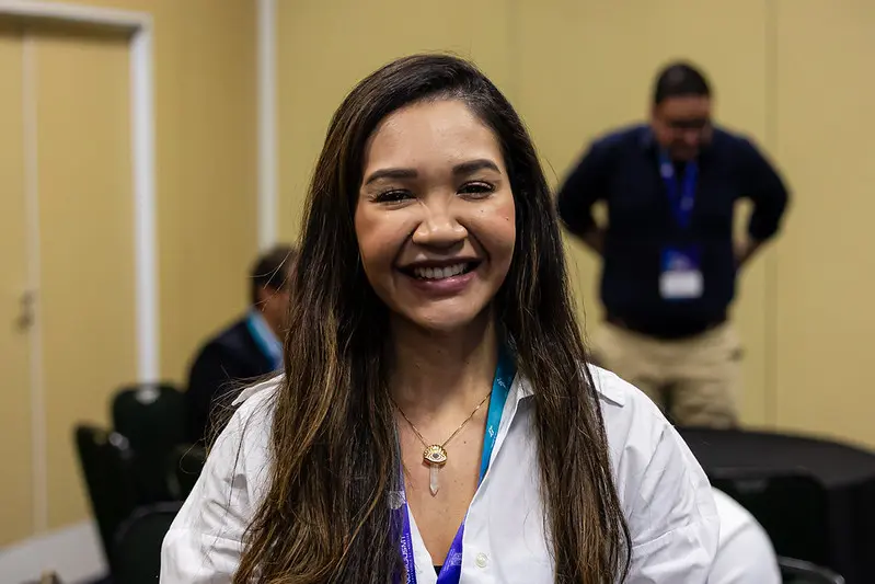 Foto horizontal que mostra Rayandria Santiago, gerente de projetos do Núcleo de Projetos de Inovação do Tribunal de Justiça de Roraima, do busto pra cima, em pé, sorrindo pra foto. Ela é uma mulher jovem, de longos cabelos castanhos e lisos, usando camisa branca.