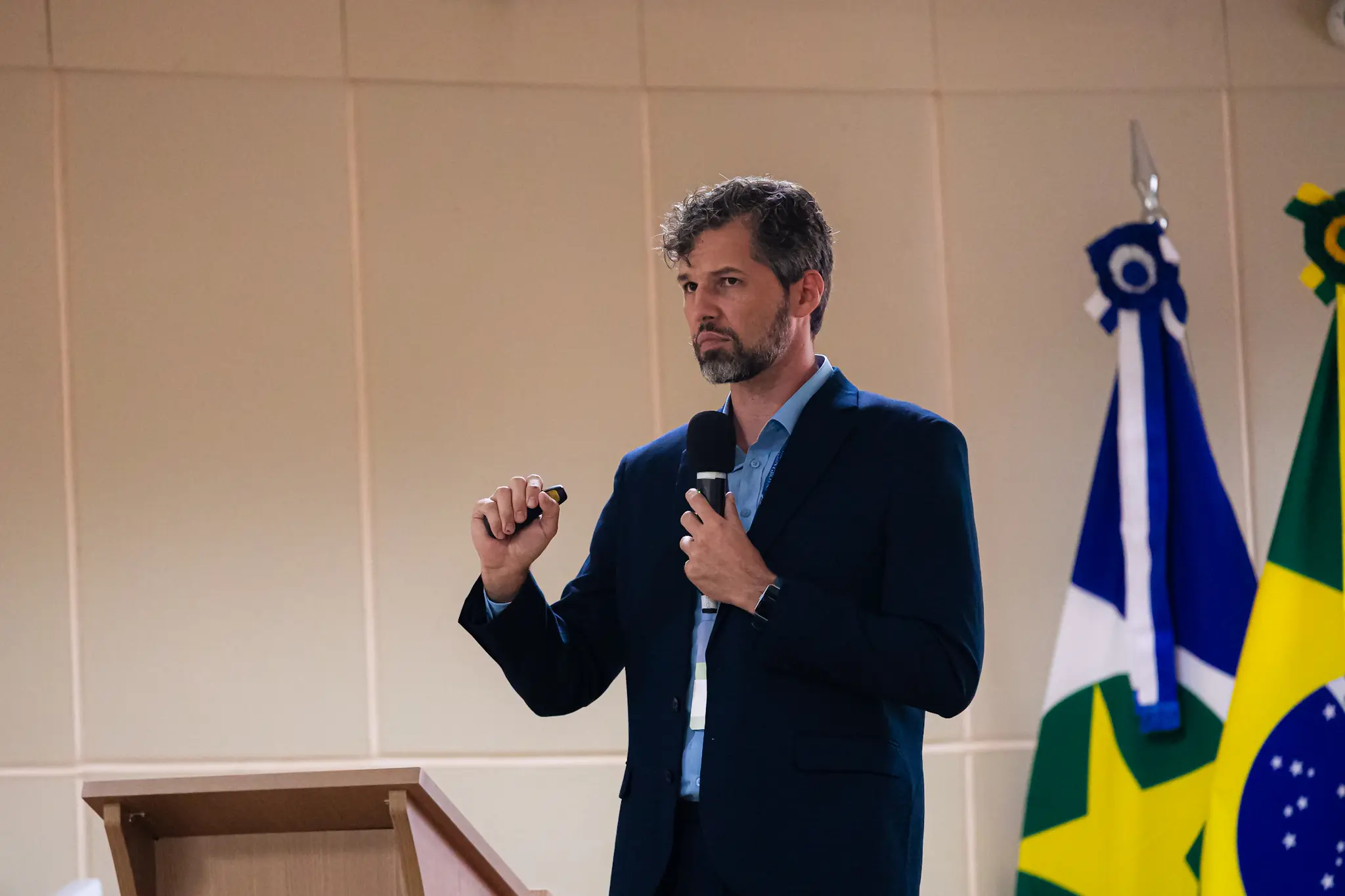Homem de cabelos grisalhos e barba, terno azul-marinho e camisa clara, discursa ao microfone com crachá institucional, bandeiras de MT e do Brasil ao fundo.