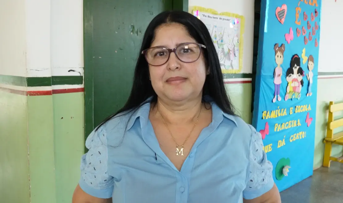 Mulher de óculos e blusa azul-clara, colar com pingente em "M", posa em corredor escolar com painel decorativo sobre família.