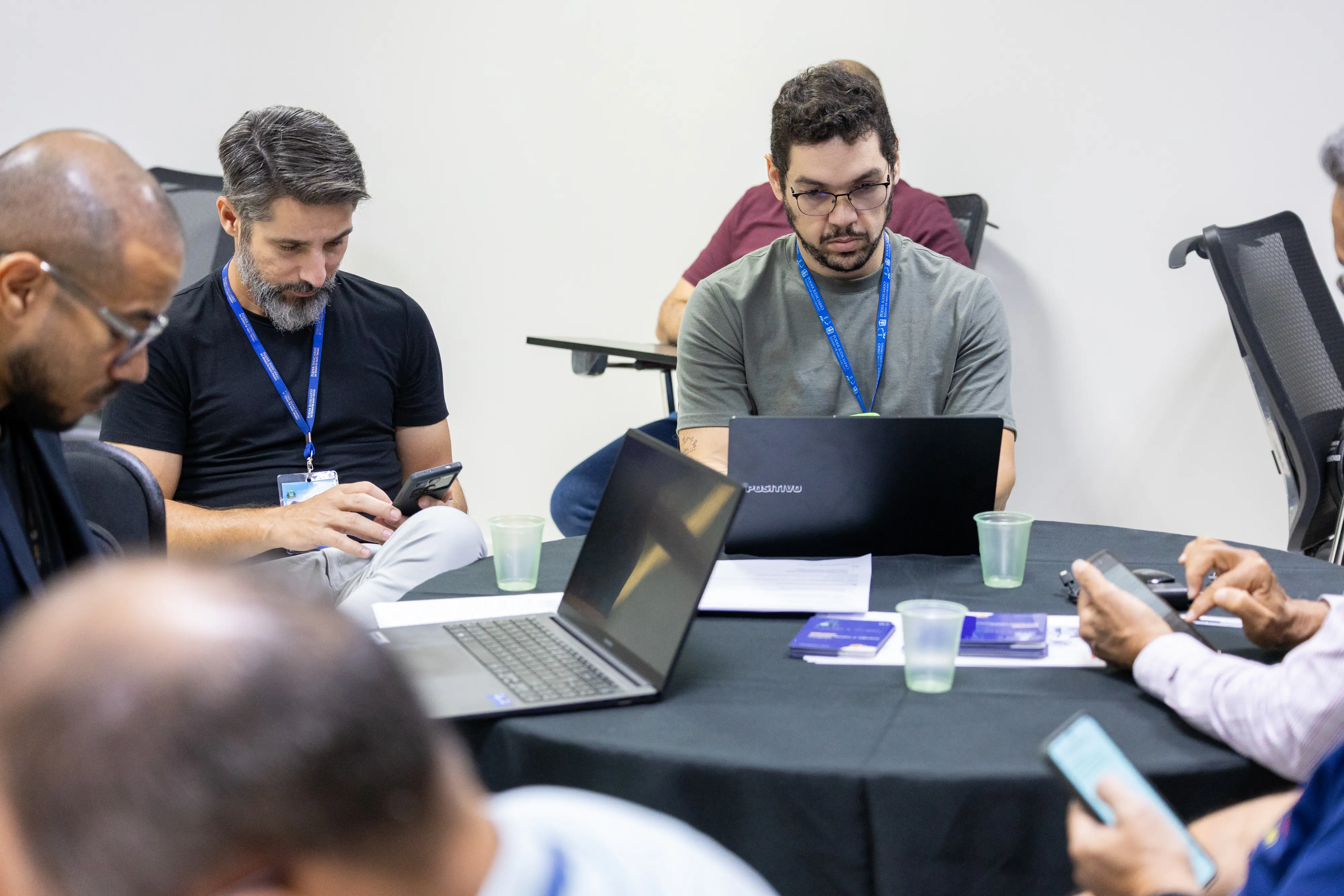 Homens com crachá institucional concentram-se em celulares e notebook. Sentados em torno da mesa, analisam documentos impressos, mantendo postura atenta durante a dinâmica prática.