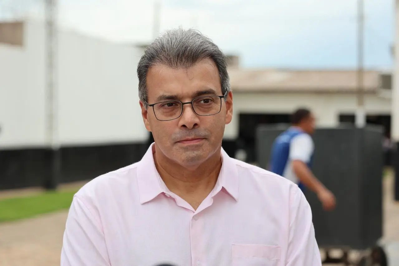 Retrato frontal de um homem de óculos e camisa rosa clara, com expressão séria. Ele está em um pátio externo de piso sextavado, com um prédio branco e gramado ao fundo sob um céu nublado.
