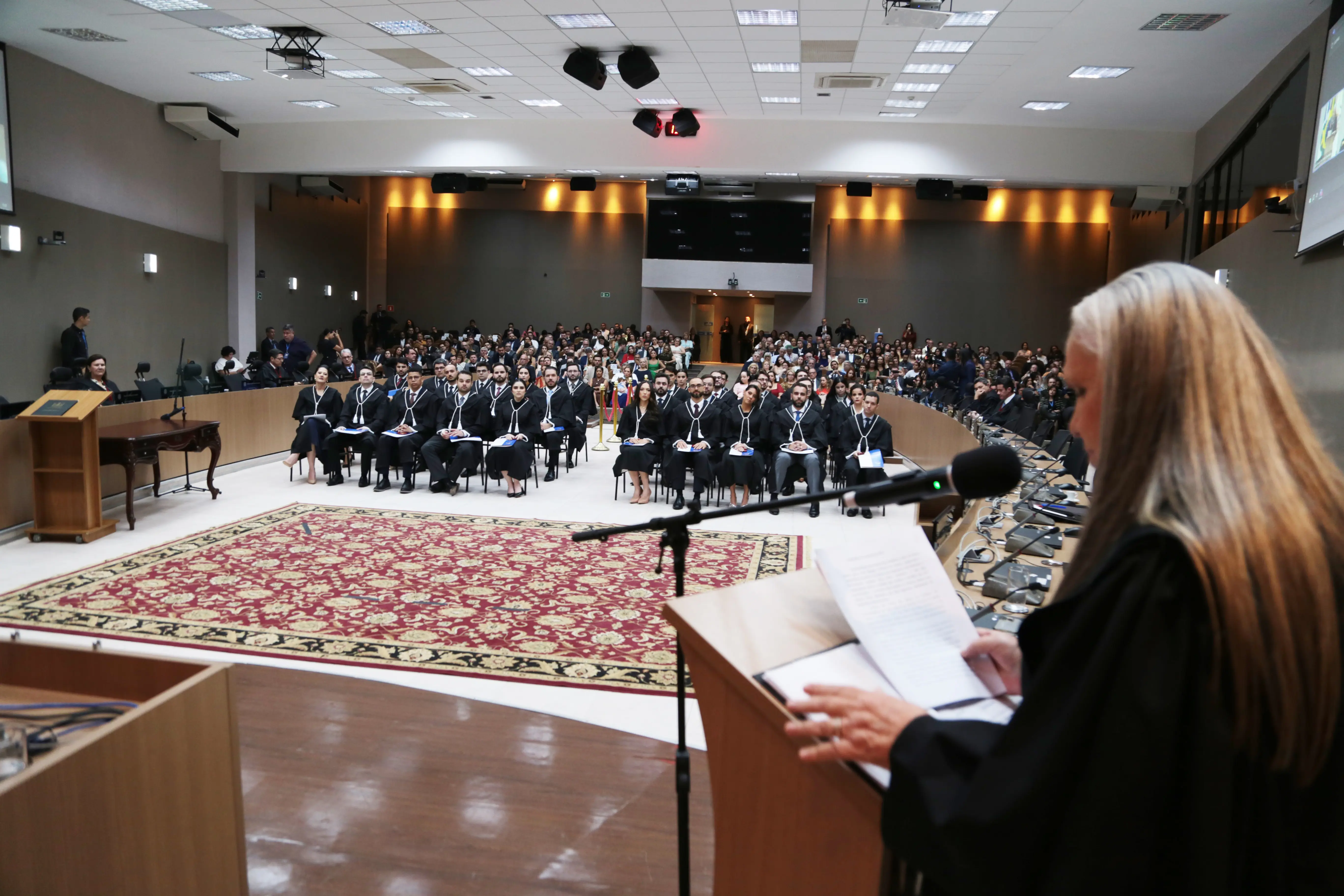 Desembargadora fala ao microfone para um auditório lotado durante solenidade. Magistrados togados sentados nas primeiras fileiras. Público acompanha cerimônia com atenção.