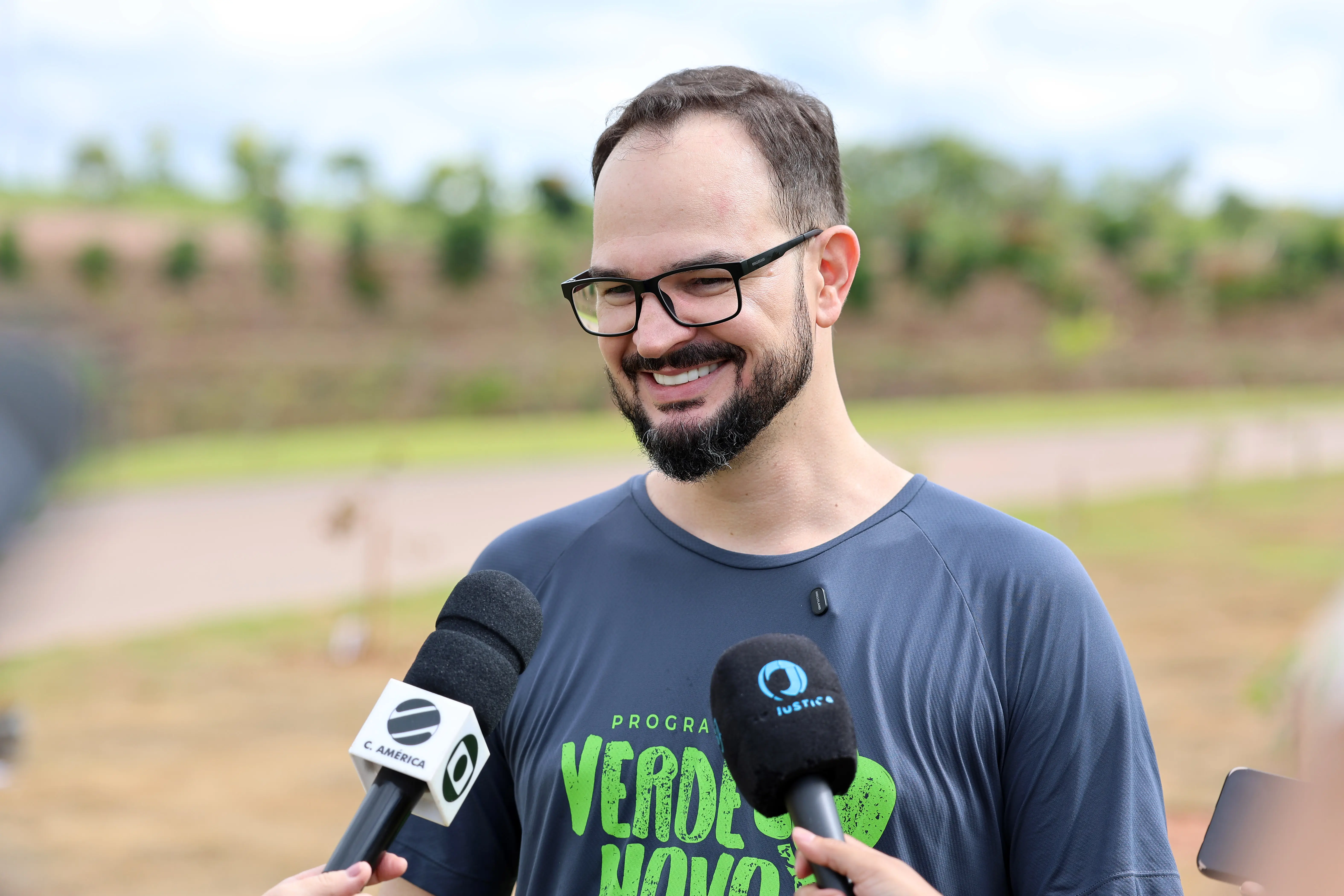 Homem de pele clara, barba curta e óculos, sorrindo durante uma entrevista ao ar livre. Veste camiseta cinza com a frase 