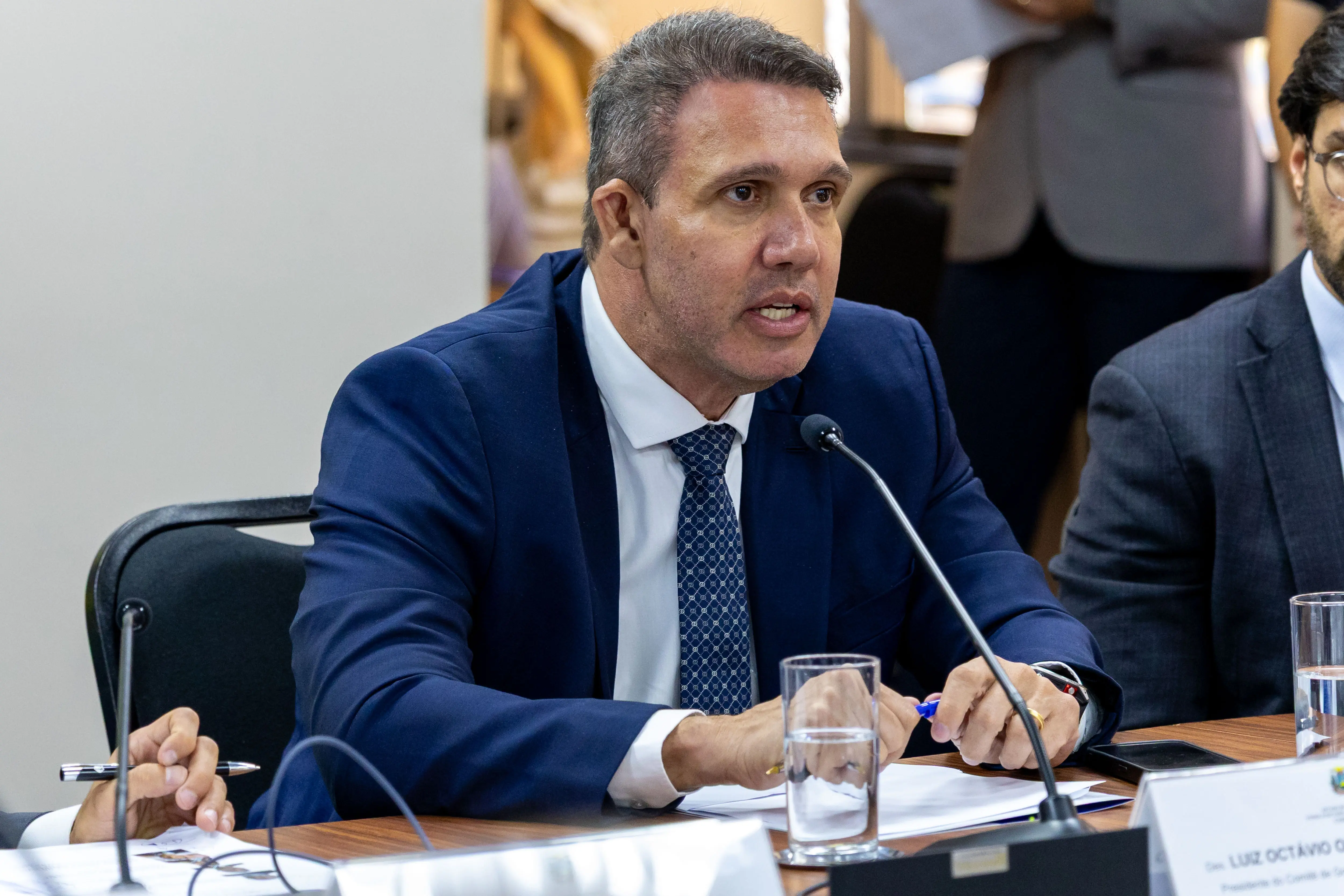 Um homem de pele clara e cabelos curtos acinzentados, vestindo paletó azul-marinho e gravata, fala ao microfone em uma reunião. Ele segura uma caneta azul e tem papéis à sua frente sobre a mesa.