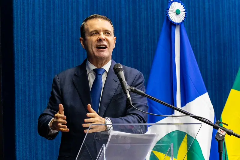 palestrante Edilson Mougenot. A imagem em plano médio registra um homem de terno azul escuro e gravata azul, gesticulando expressivamente enquanto discursa em um púlpito de acrílico. 
