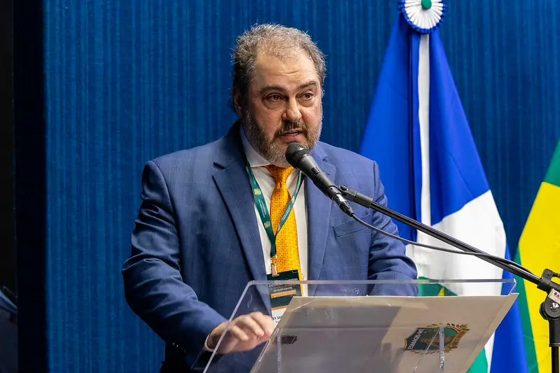 Desembargador Wesley Lacerda. A imagem em plano médio registra um homem barbado, de terno azul e gravata laranja/amarela, discursando em um púlpito de acrílico. Ele usa um crachá pendurado em uma fita amarela e fala em um microfone ajustável. No púlpito, é visível um papel