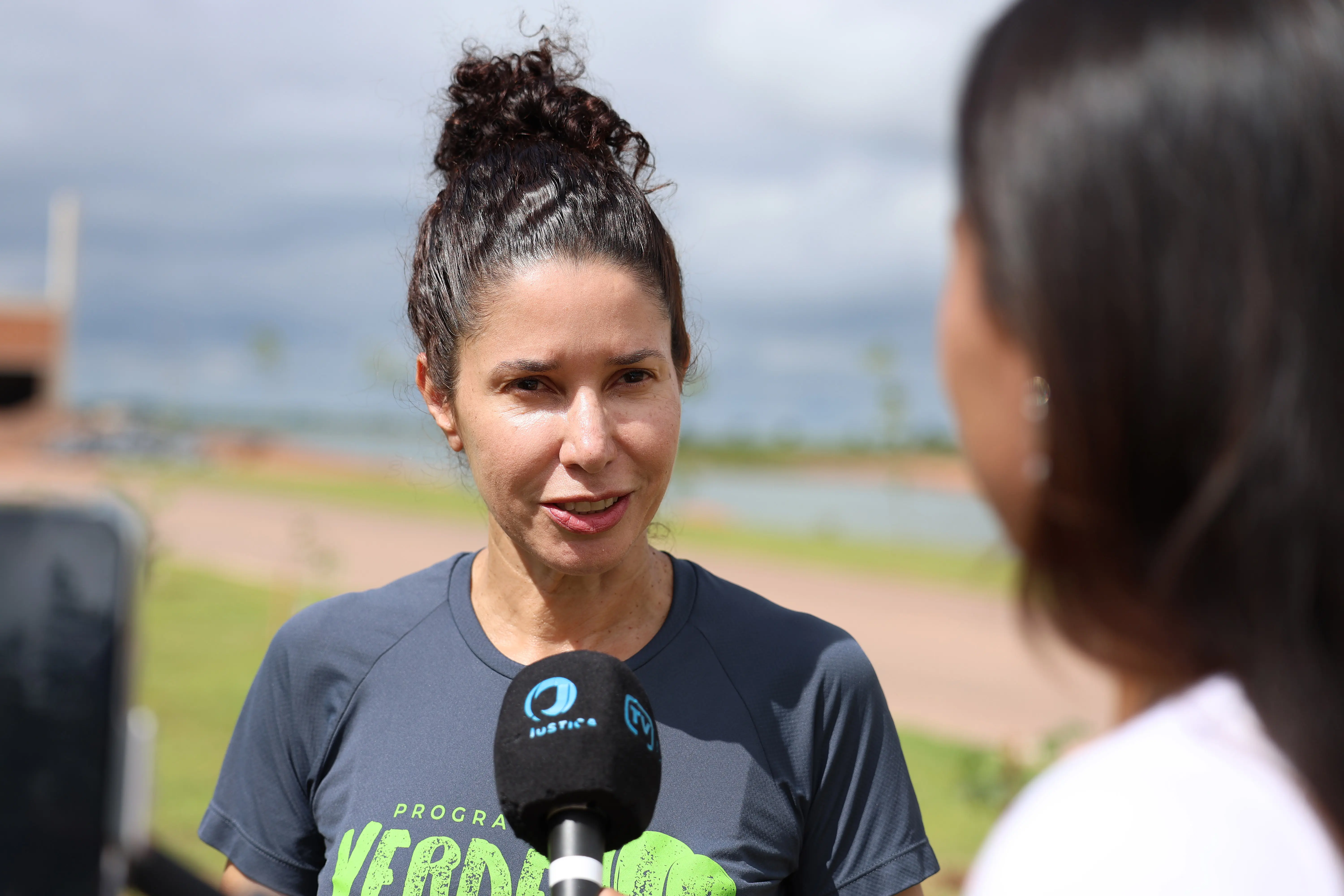  Mulher de pele clara e cabelos escuros presos em um coque, sorrindo durante entrevista. Veste camiseta cinza do 