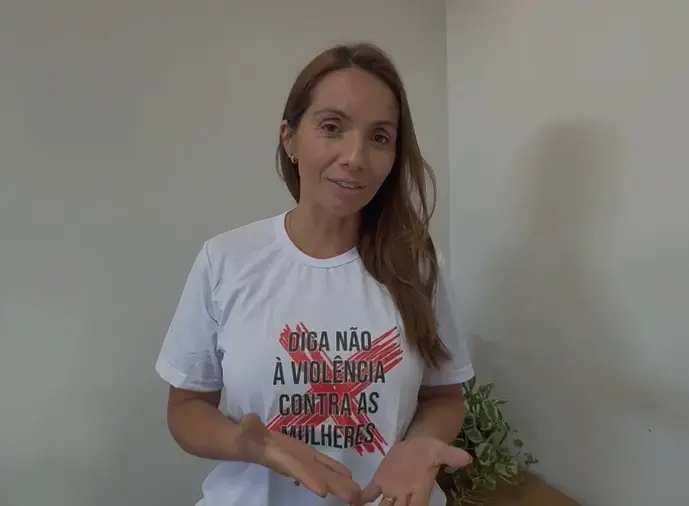 Foto horizontal colorida que mostra a vereadora de Tapurah, Daniele Zottis, em pé. Ela é uma mulher branca, magra, de cabelos loiros, longos, lisos e soltos, usando camiseta branca com a estampa de um X vermelho e a frase: Diga não à violência contra as mulheres