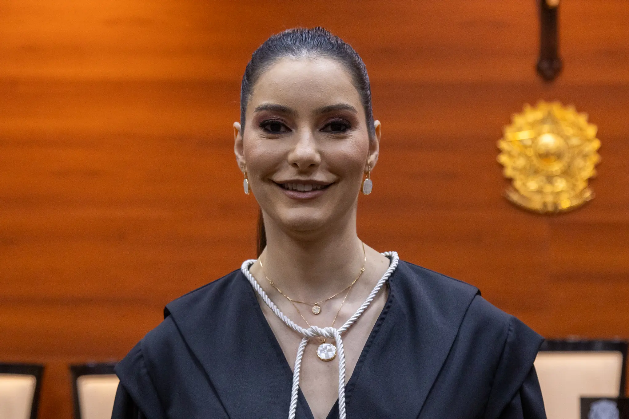 Foto horizontal colorida que mostra a juíza Patrícia Bedin, do busto pra cima, posando sorrindo pra foto, em pé, usando toga, no Plenário do Tribunal de Jsutiça. Ela é uma mulher branca, magra, alta, de cabelos lisos e pretos, presos em rabo-de-cavalo, e maquiada.