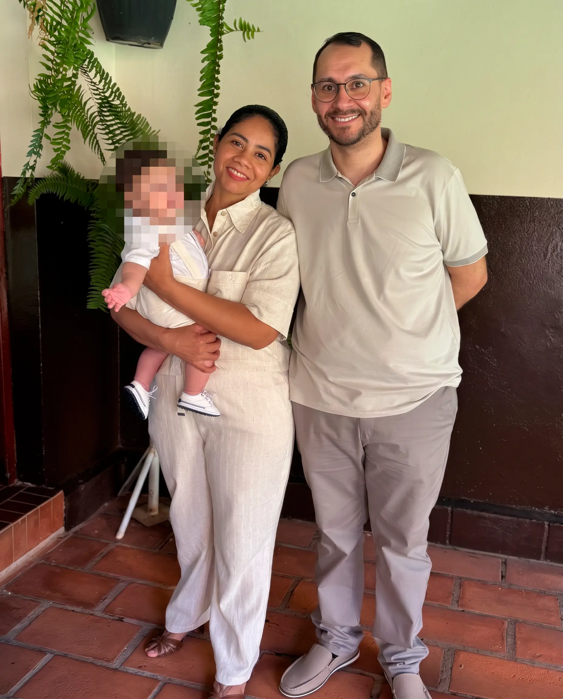 Homem de óculos e camisa polo clara ao lado da esposa, que usa conjunto de linho claro. Ela segura no colo um bebê de roupa branca. A criança tem o rosto borrado para preservação da imagem. Eles estão em pé sobre piso de lajotas escuras com planta ao fundo.