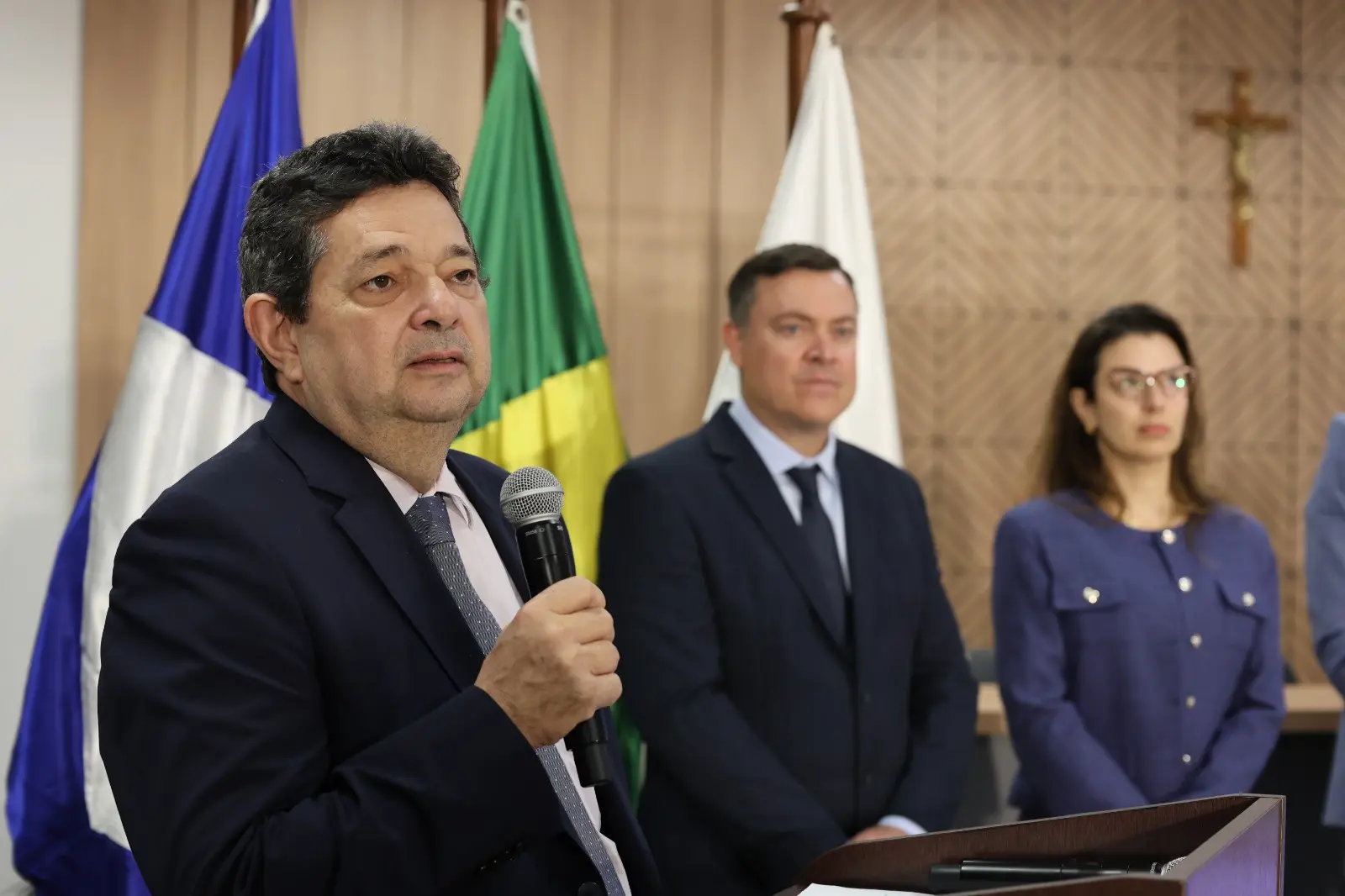 O corregedor des. José Luiz Leite Lindote, segura um microfone e usa terno escuro com gravata cinza. Ao fundo aparecem as bandeiras e autoridades que participaram da solenidade.
