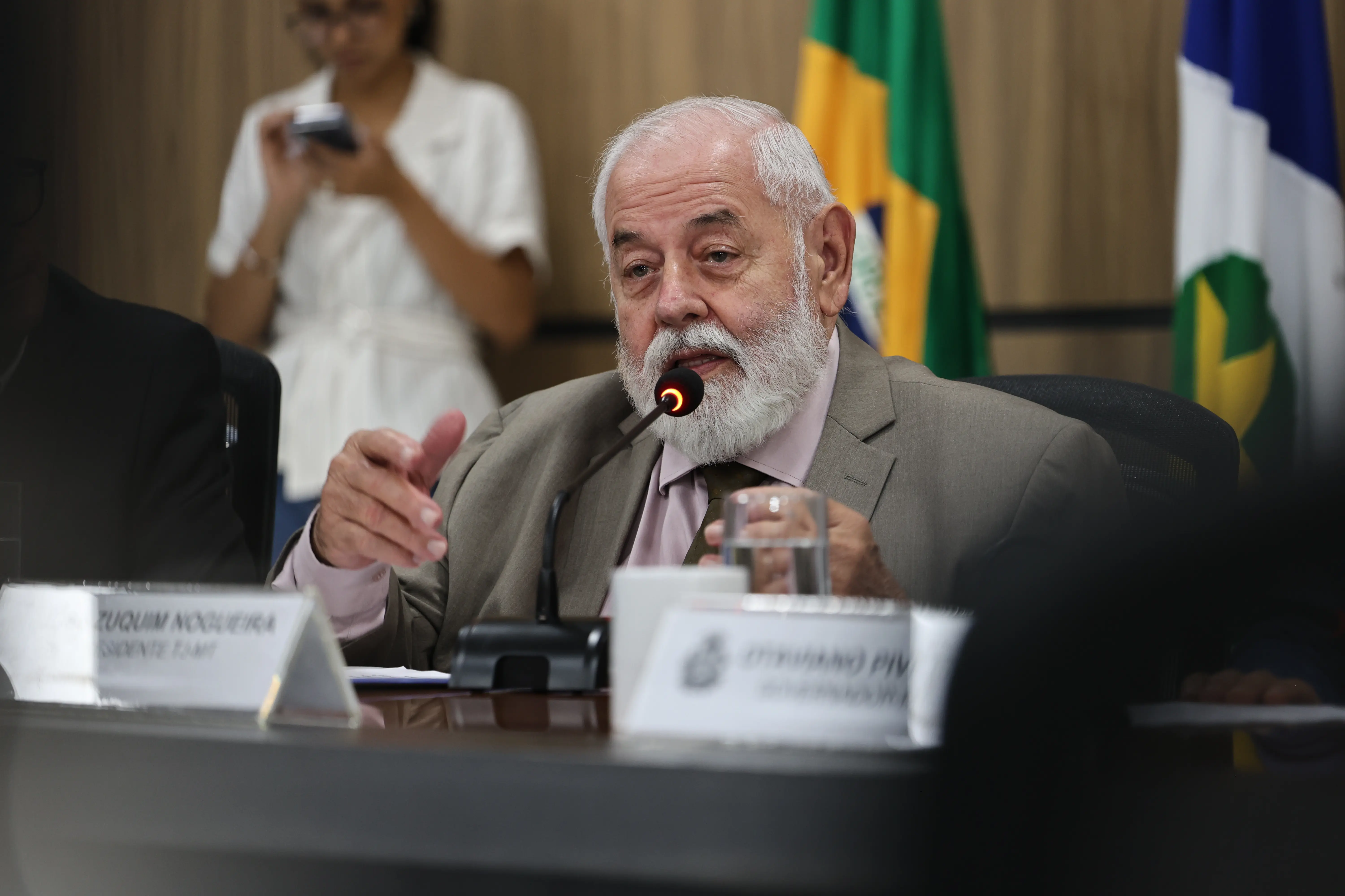 Homem idoso, branco, de barba e cabelos grisalhos, veste terno cinza e fala ao microfone integrado à mesa. Ele gesticula com a mão direita. Ao fundo, uma pessoa desfocada e bandeiras oficiais.
