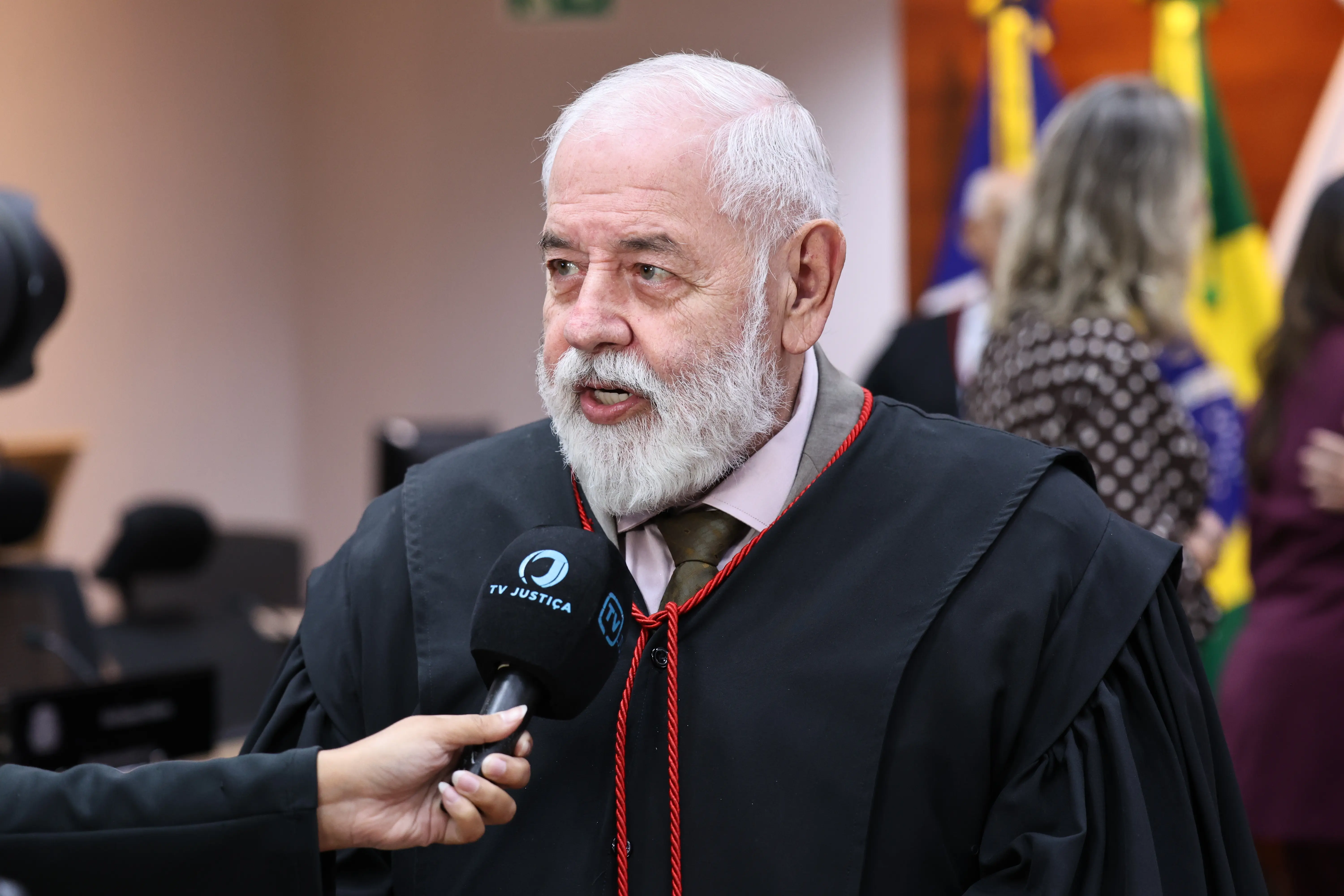 Homem idoso de barba branca e toga preta concede entrevista a um microfone com a marca 