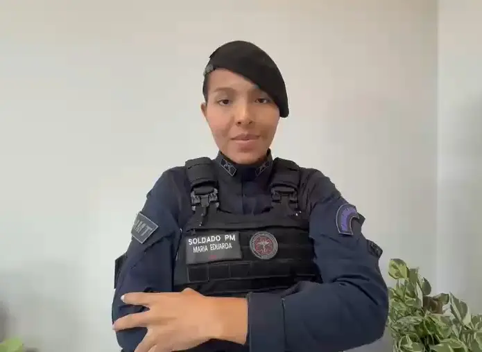 Foto horizontal colorida que mostra a soldado PM Maria Eduarda, da cintura pra cima, em pé, de braços cruzados. Ela é uma jovem, negra, de olhos escuros, usando farda e boina preta. 