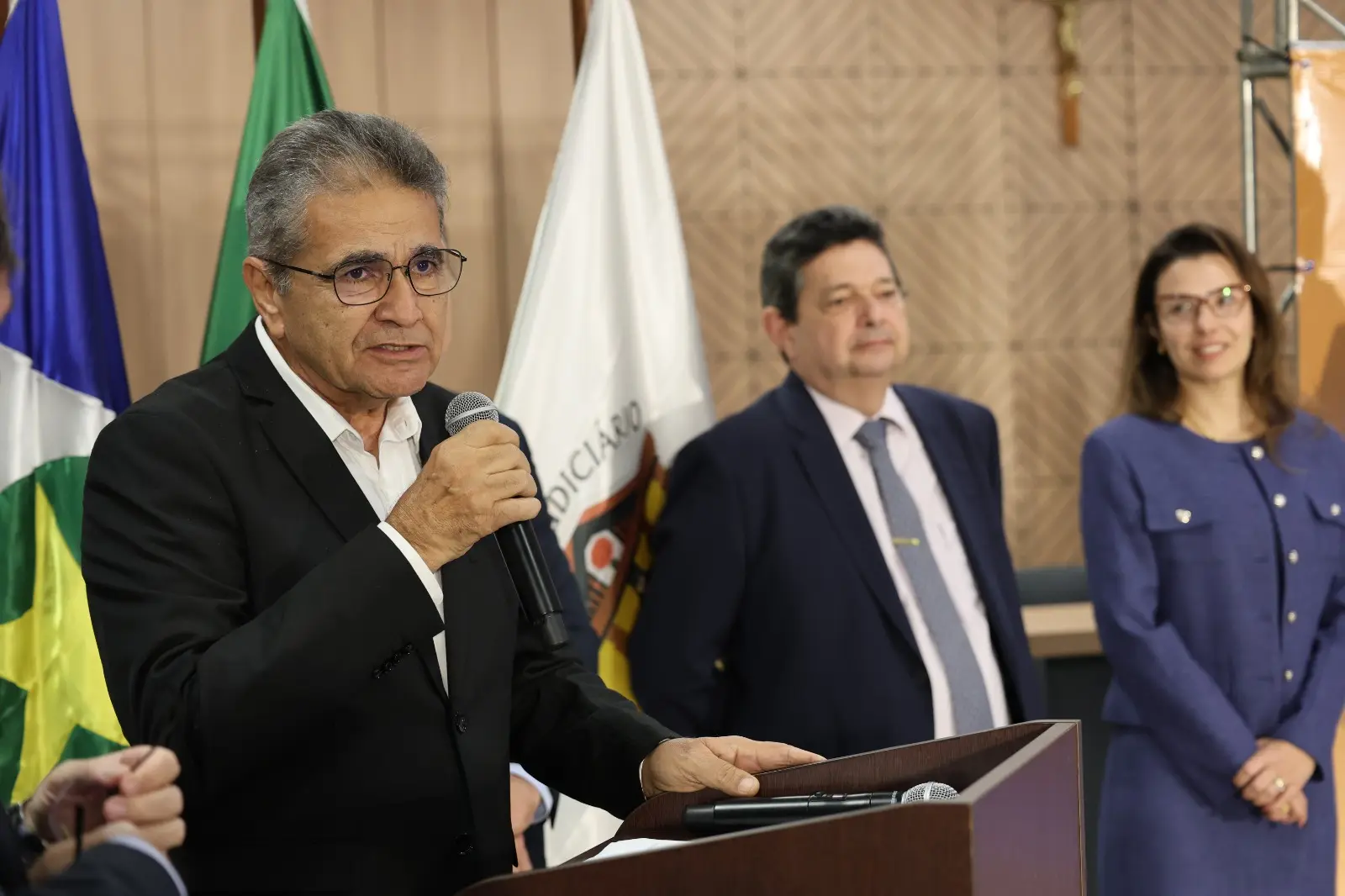Presidente do Intermat, Francisco Serafim, segura um microfone durante pronunciamento na cerimônia de entrega de títulos. Ao fundo aparecem o corregedor, desembargador José Luiz Leite Lindote, e a juíza auxiliar da Corregedoria, Myrian Pavan Schenkel, acompanhando a solenidade.