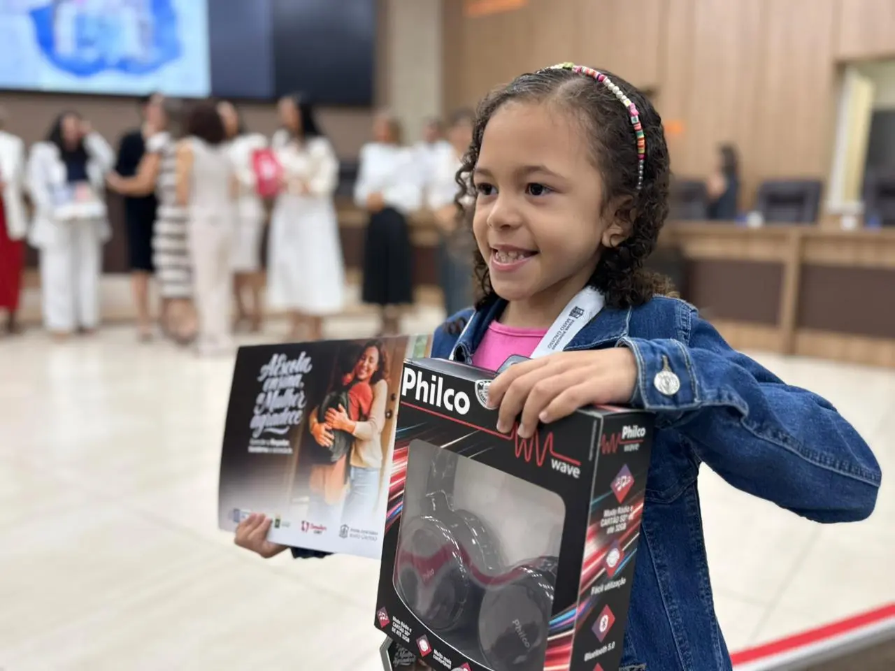 Em primeiro plano, uma menina sorridente com jaqueta jeans segura a caixa de um fone de ouvido Philco e um cartaz do projeto. Ao fundo, pessoas conversam dispersas em um grande salão.