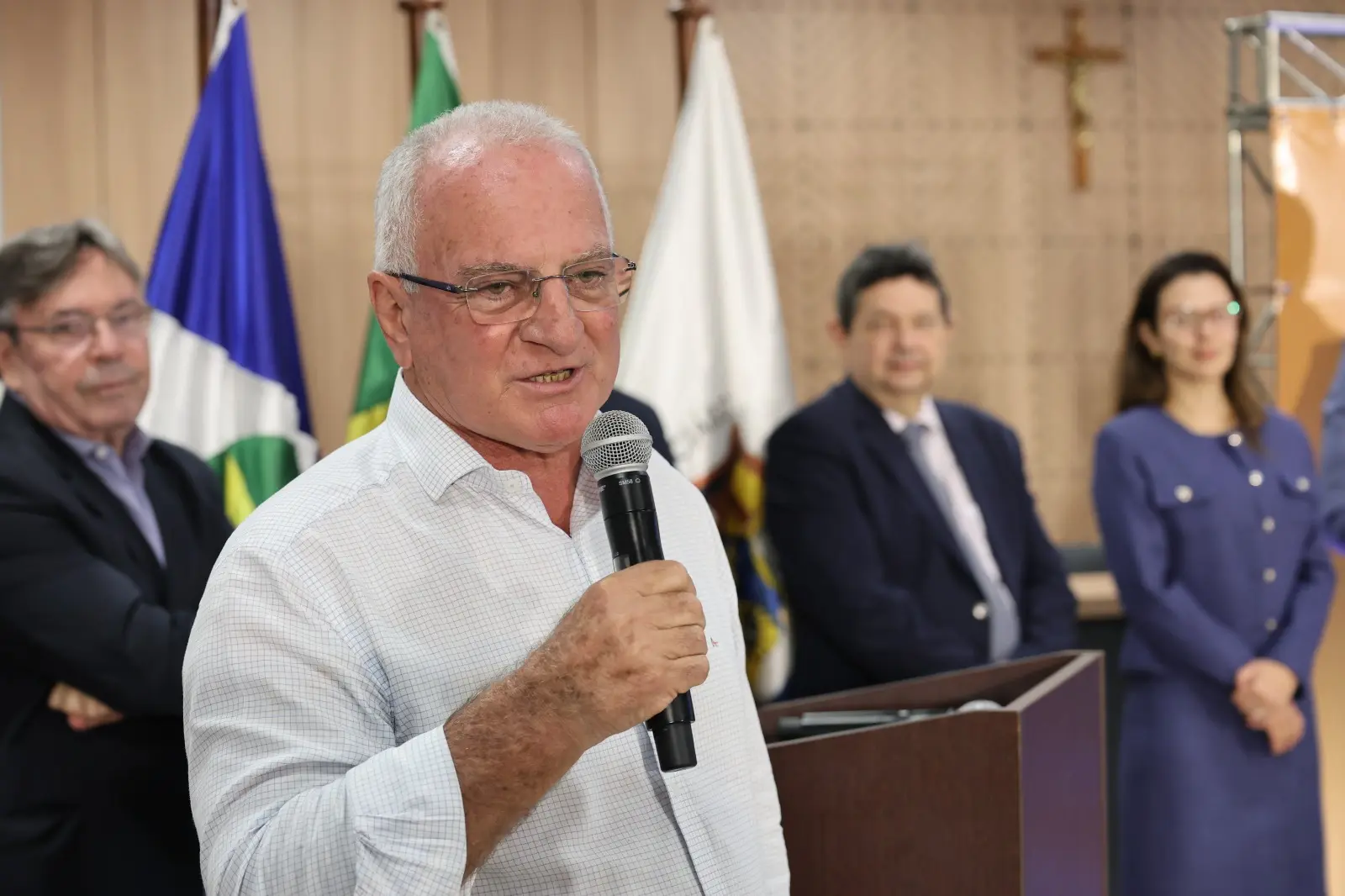Prefeito de Chapada dos Guimarães, Osmar Froner, segura um microfone durante pronunciamento na cerimônia de entrega de títulos. Ao fundo aparecem o corregedor, desembargador José Luiz Leite Lindote, e a juíza auxiliar da Corregedoria, Myrian Pavan Schenkel.