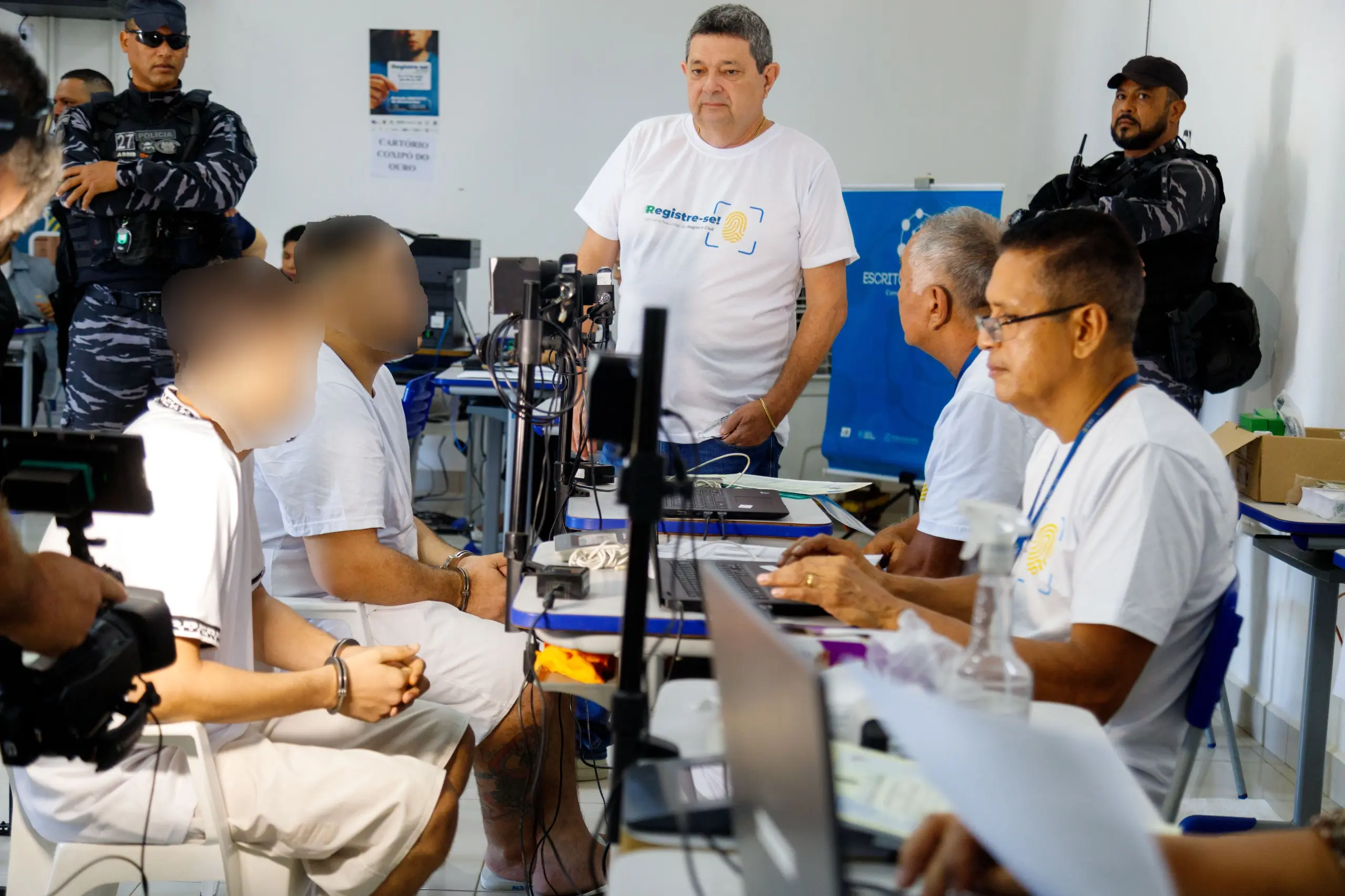 Dois reeducandos, com roupas brancas e algemas nos pulsos, estão sentados frente a frente com servidores que realizam atendimento em computadores, no mutirão do programa “Registre-se!” dentro da Penitenciária Central do Estado (PCE).