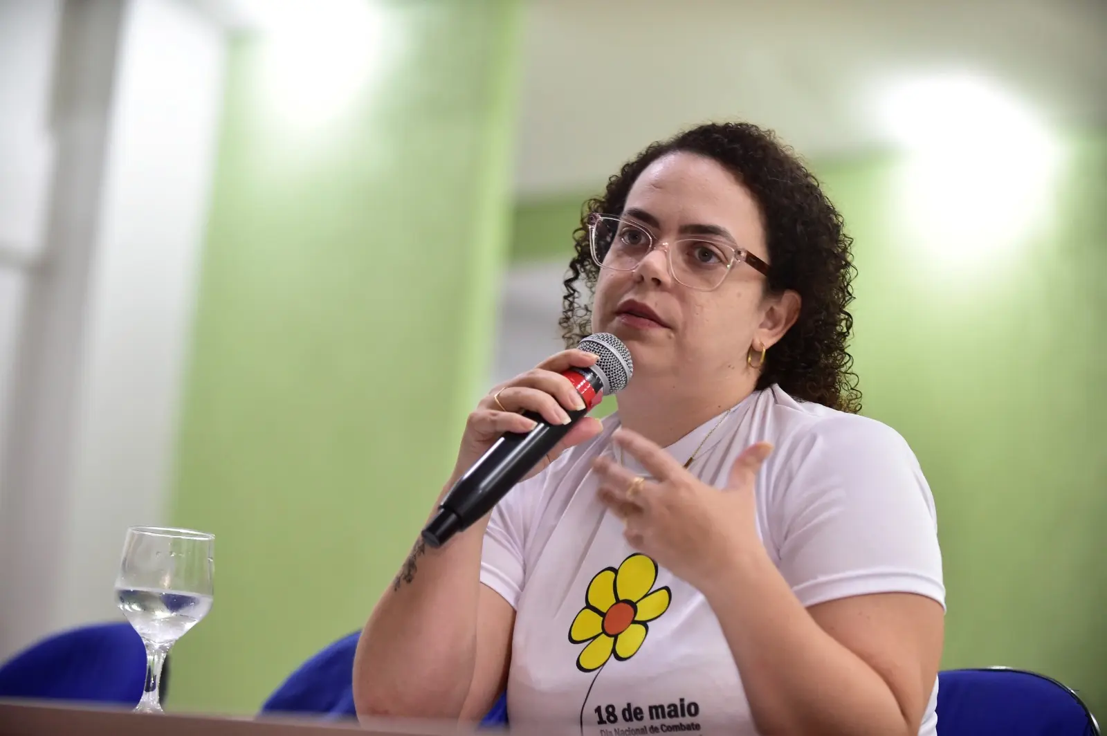 Ana Letícia Bonfanti é uma mulher morena, de cabelos cacheados e escuros, usa óculos de grau e camiseta branca