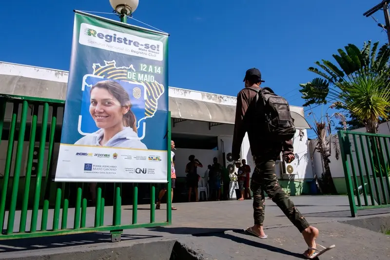  Foto do banner da campanha “Registre-se! Mato Grosso – Semana Nacional do Registro Civil” afixado em grade verde na entrada de um prédio público em Cuiabá. O cartaz traz a imagem de uma mulher sorridente com fundo azul e o mapa do Brasil em destaque, com datas de atendimento entre 12 a 16 de maio. 