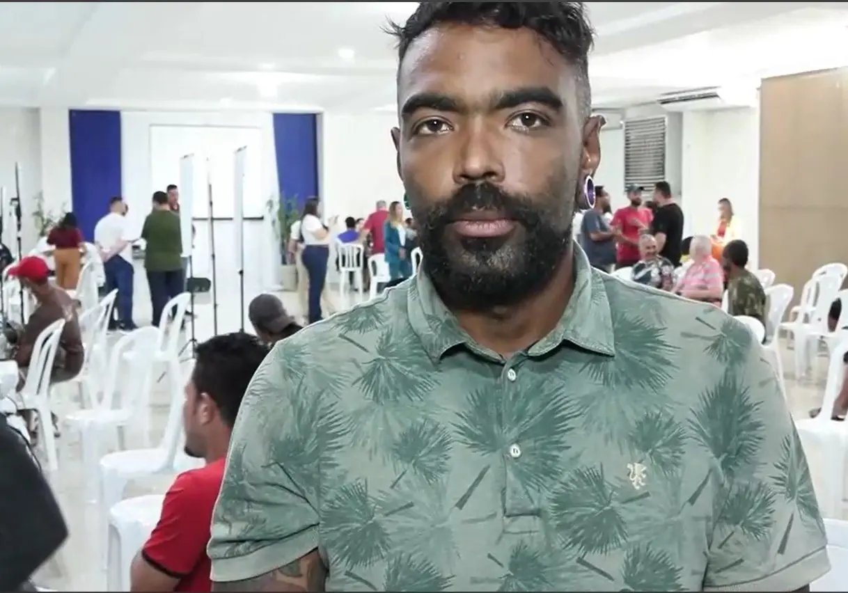 Foto de homem negro com barba e cabelos curtos aparece em destaque na imagem, olhando diretamente para a câmera. Ele veste uma camisa polo verde com estampa de folhas e tem alargador na orelha. Ao fundo, várias pessoas estão sentadas ou em pé em um salão bem iluminado, com cadeiras brancas.