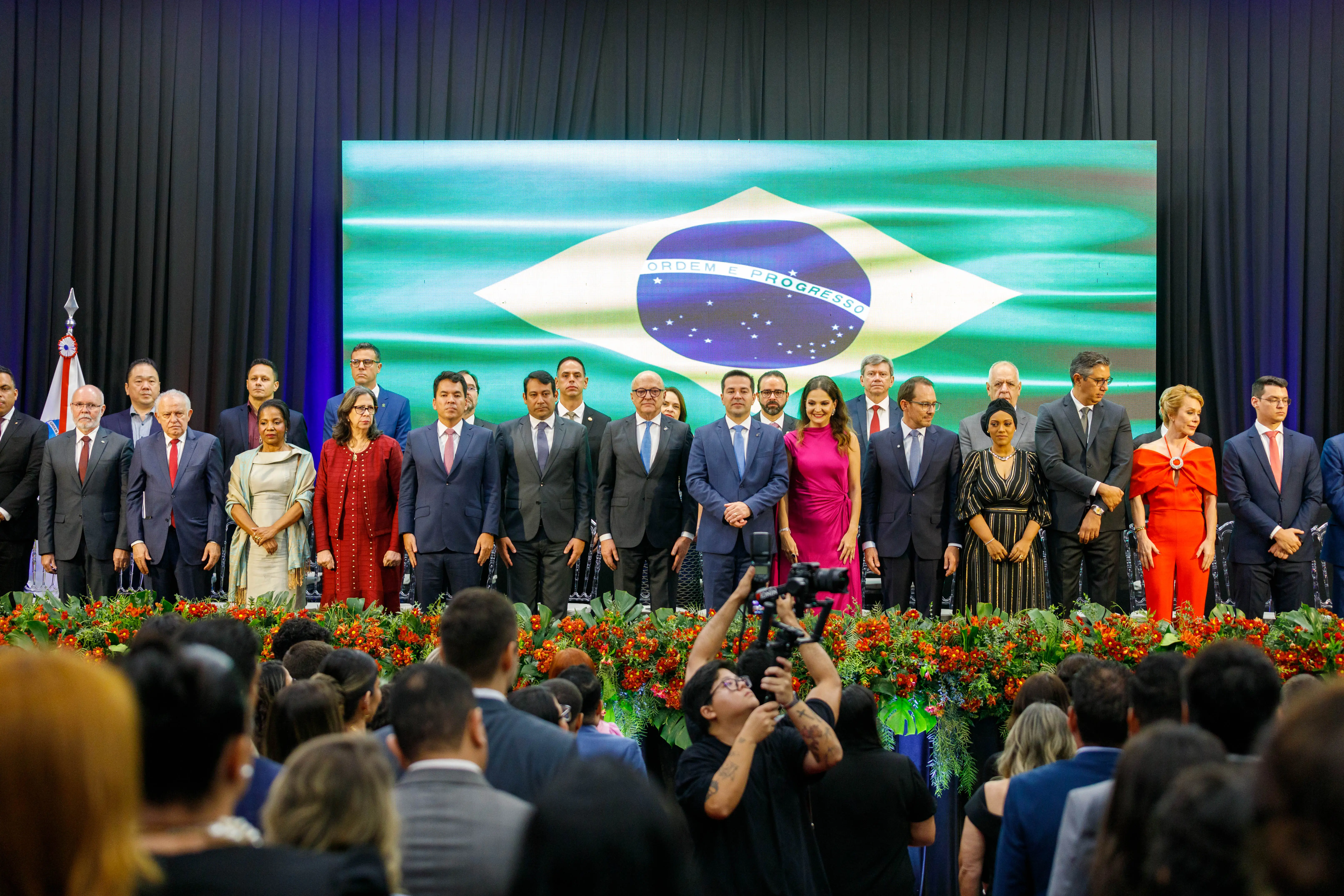 A imagem mostra um grupo de pessoas em pé em um palco em frente a uma grande bandeira brasileira. O dispositivo é formado por seis mulheres e 20 homens, todos vestidos com trajes formais. Em frente ao palco, há uma fileira de arranjos florais vermelhos e verdes.