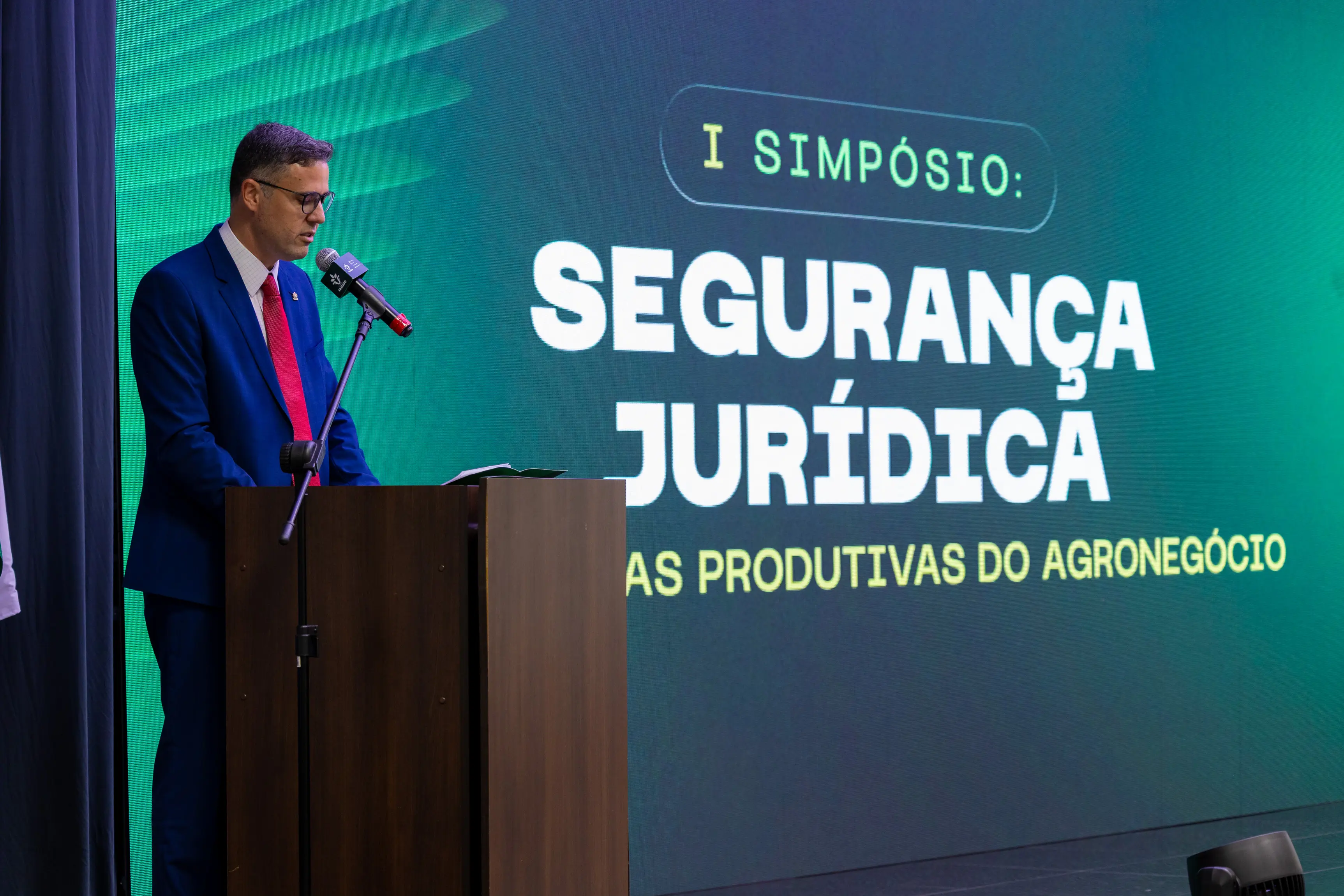 Um homem de cabelos escuros, óculos, veste terno azul e gravata vermelha, discursa em microfone sobre púlpito de madeira. Ao fundo, telão exibe "I Simpósio: Segurança Jurídica".