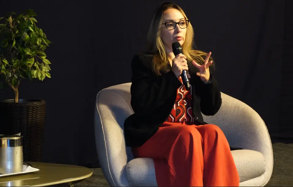 Mulher loira de óculos, blazer preto e calça vermelha sentada em poltrona cinza. Ela segura um microfone e gesticula enquanto fala. Ao fundo, uma planta e parede escura.