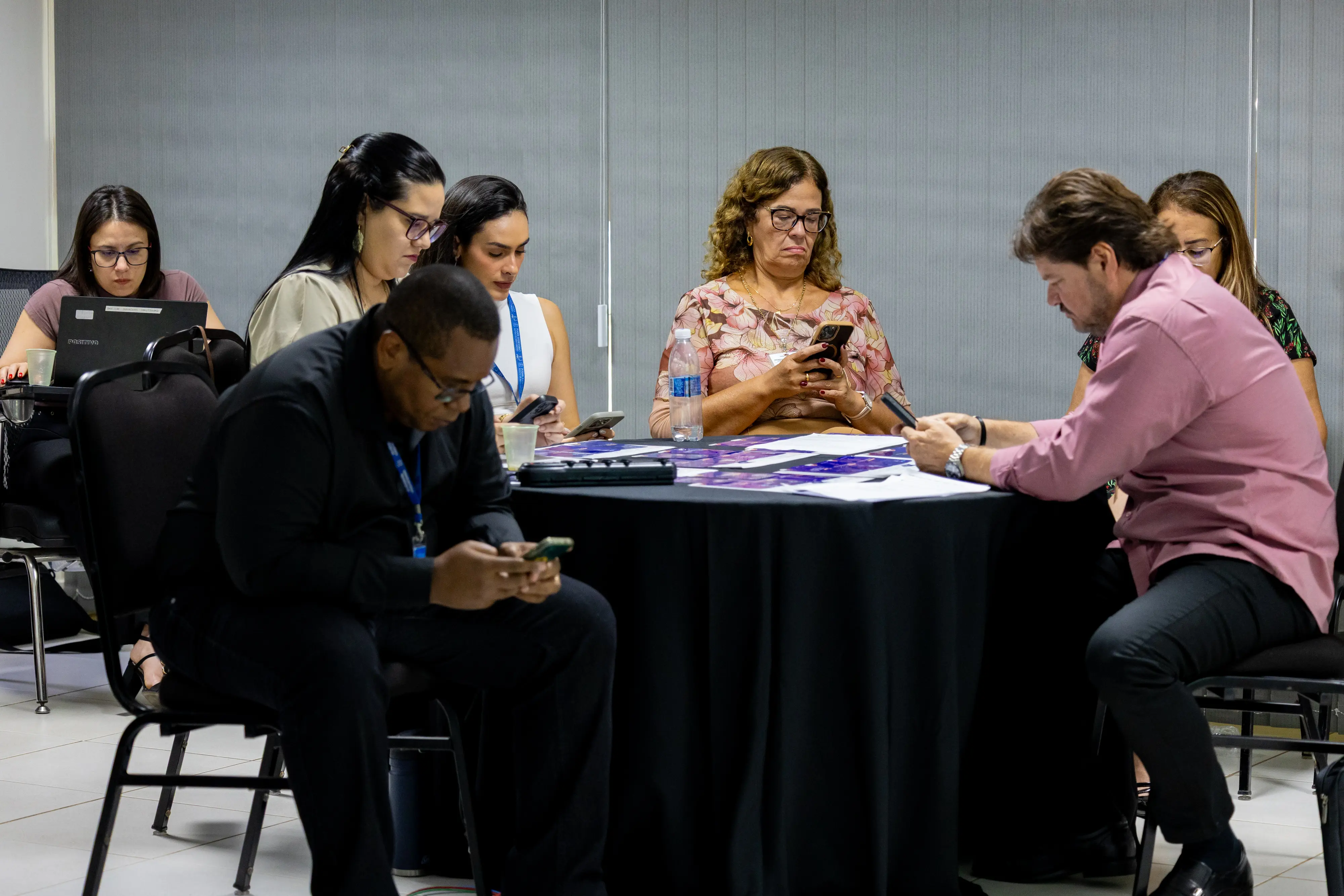 Mesa com homens e mulheres focados nos aparelhos móveis. Expressões sérias indicam concentração na tarefa proposta. Ao fundo, outros grupos repetem a mesma dinâmica.
