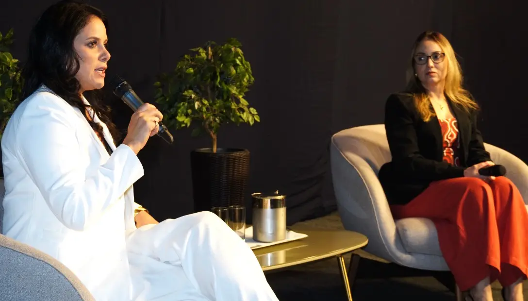 Duas mulheres sentadas em poltronas cinzas durante um evento. À esquerda, uma mulher de terno branco fala ao microfone. Ao fundo, uma planta decorativa e uma parede escura.
