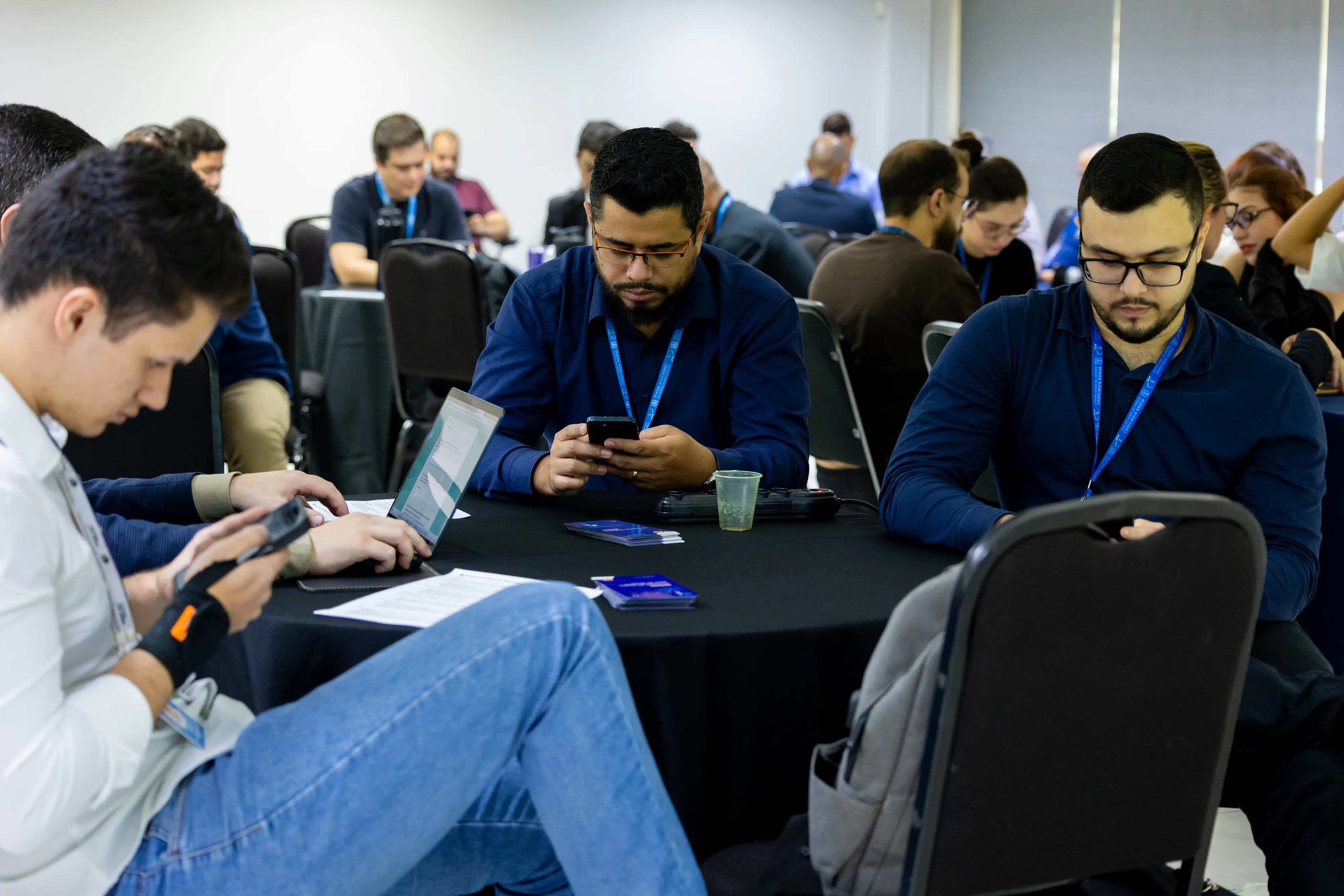 Participantes utilizam notebook e celular simultaneamente. Homem digita enquanto colegas verificam informações no telefone. Cartões e copos plásticos compõem o cenário da atividade.